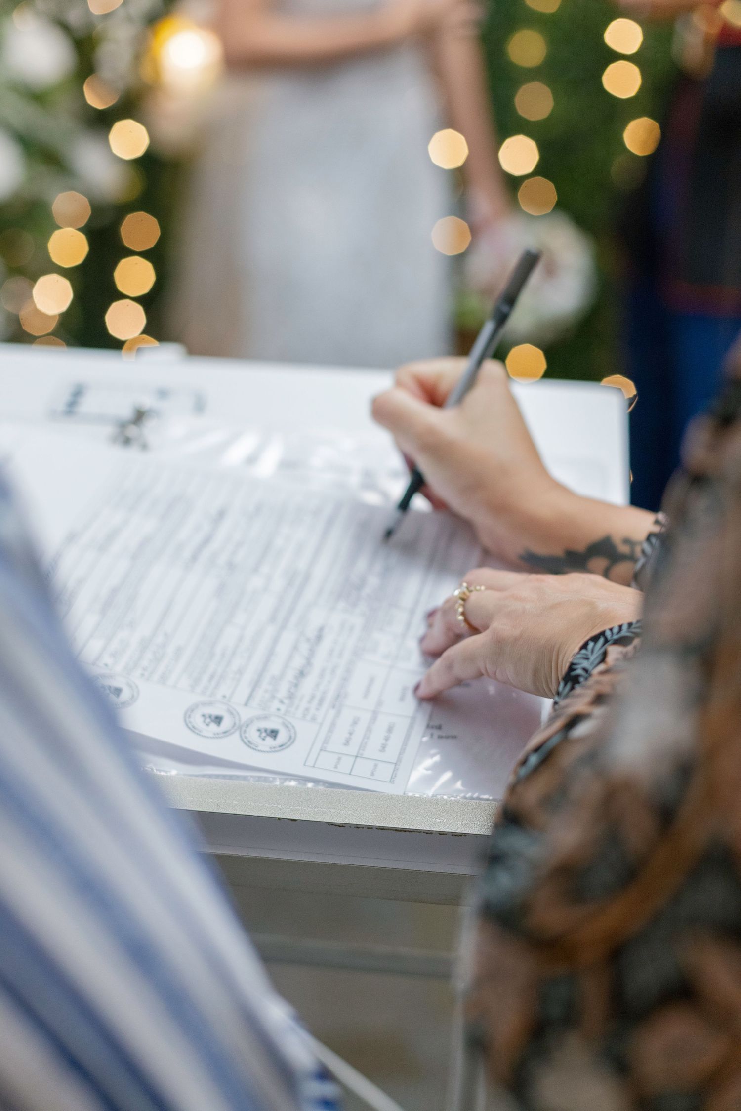 Person signing a wedding certificate at a decorated table with bokeh lights in the background.