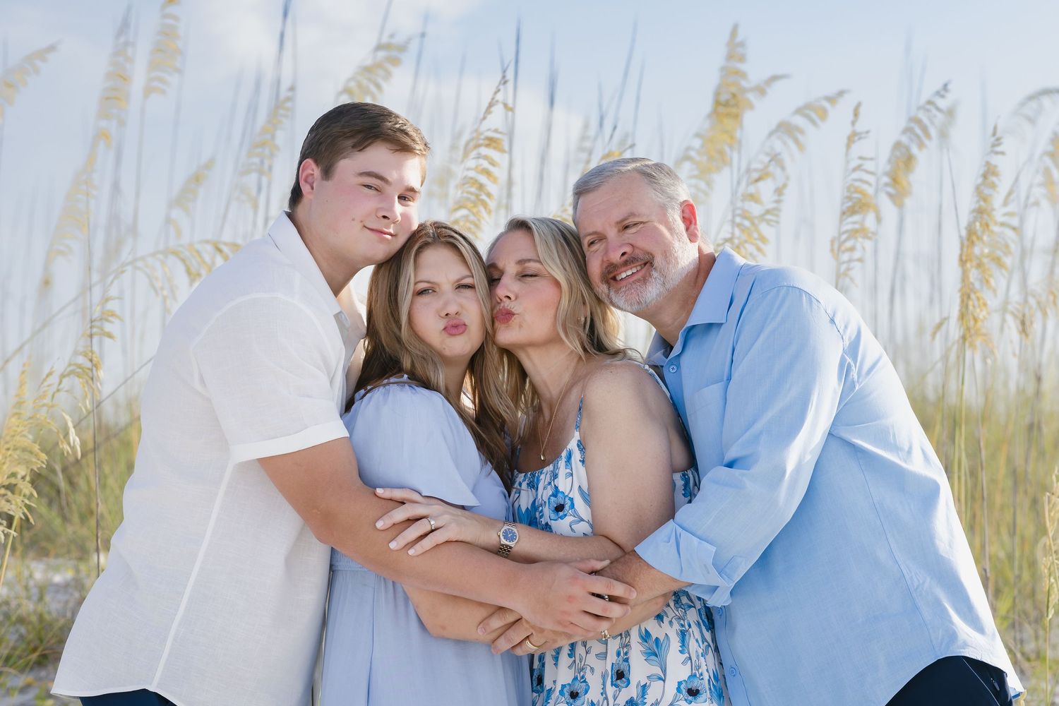 Family of four poses together on a beach with sea oats, sharing hugs and kisses in coordinating blue outfits.