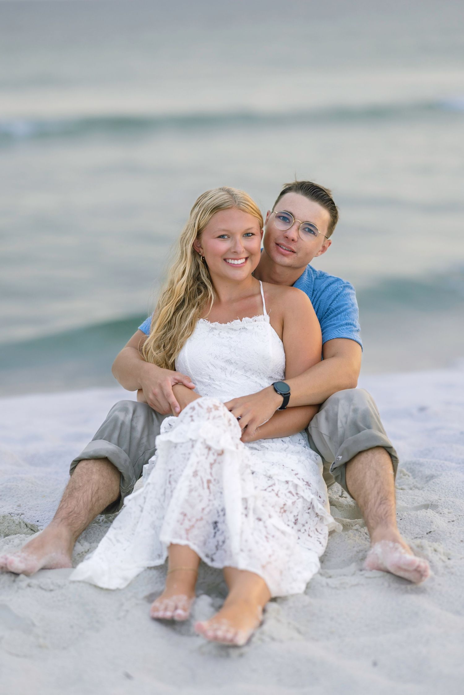 Couple sitting together on sandy beach, woman in white lace dress, man in blue shirt, smiling with ocean waves behind them.