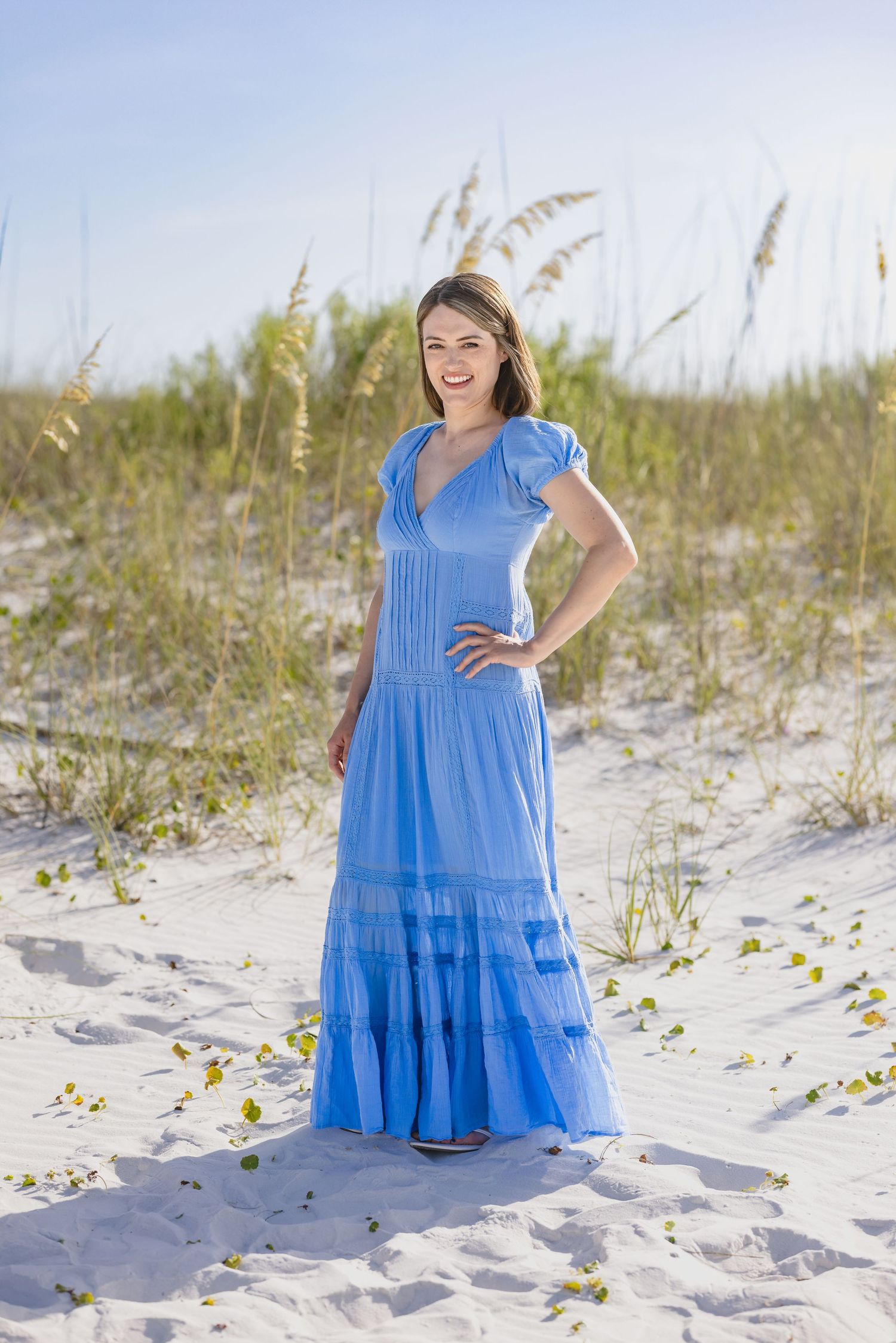 A smiling woman in a blue maxi dress poses on white sand dunes with sea oats in the background.