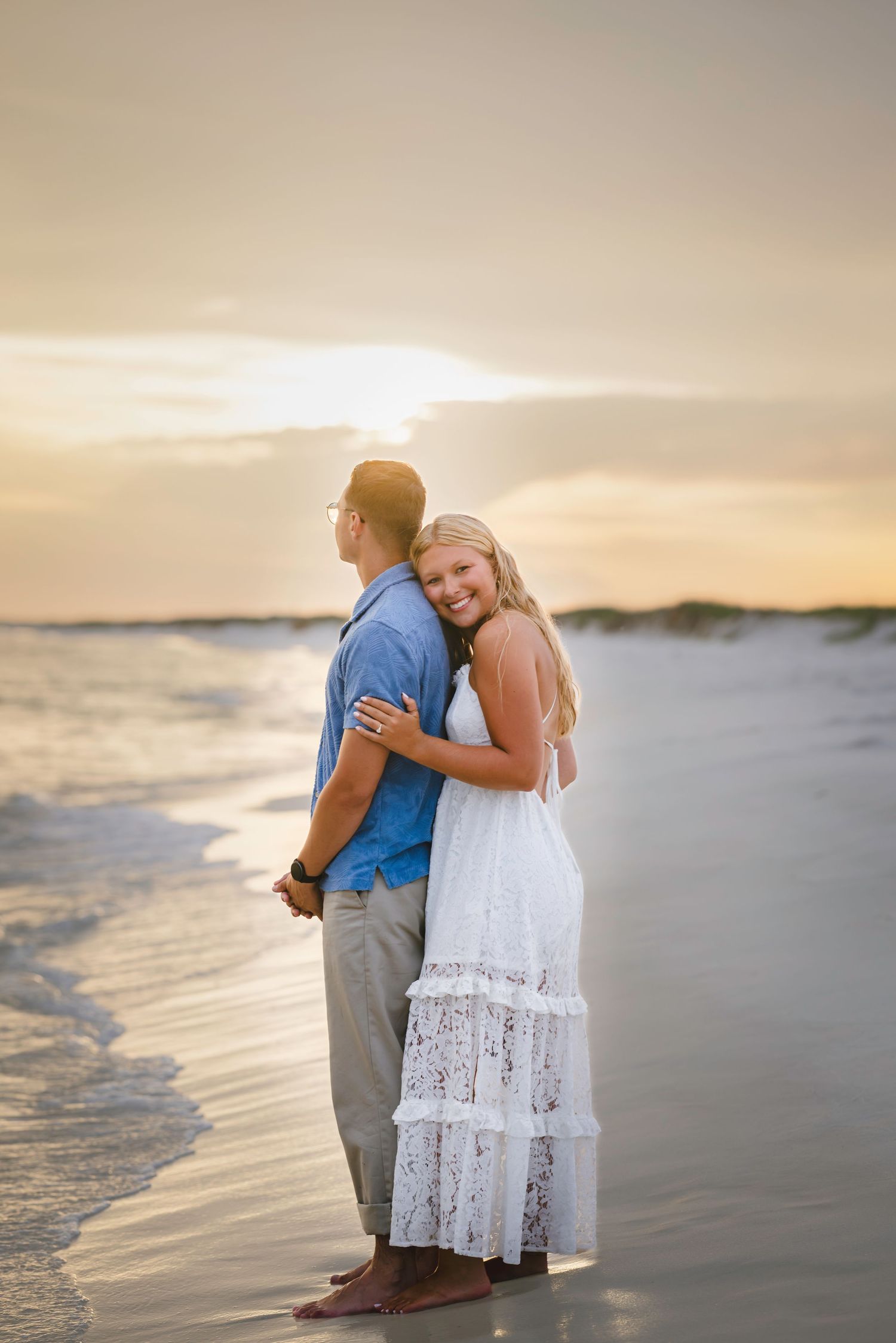 Couple embracing at sunset on a beach, woman in white lace dress smiling over her shoulder.