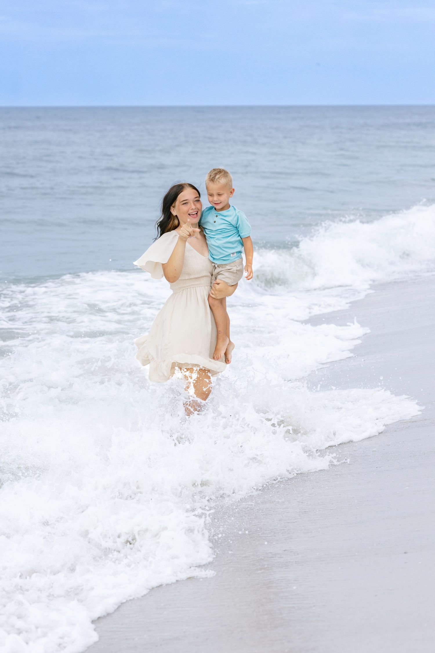 A joyful mother in a white dress carries her toddler son while running through ocean waves on a sunny beach.