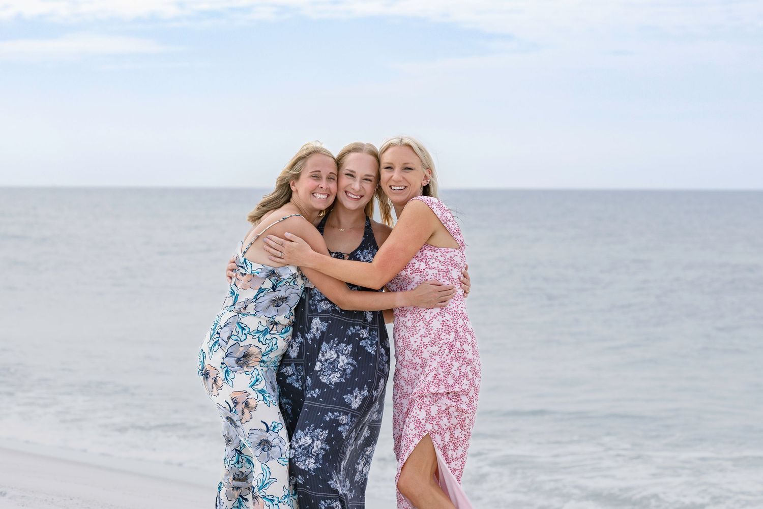 Three smiling women in floral dresses hugging on a sandy beach with ocean waves in the background.