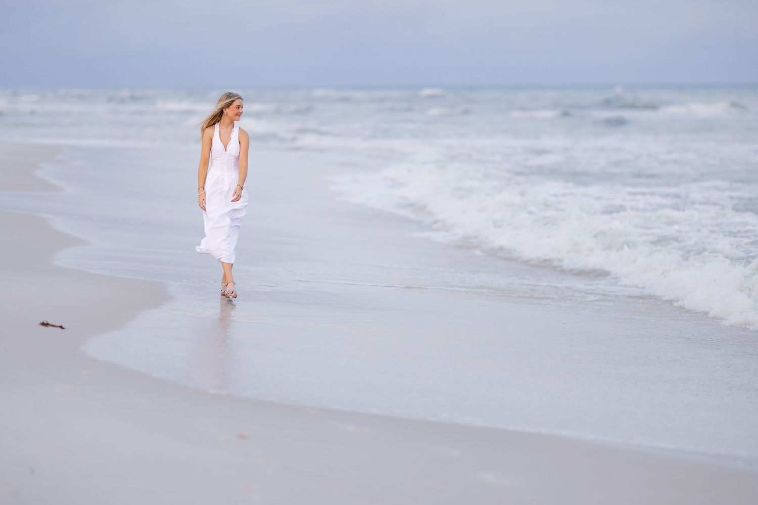 A woman in a white dress walks along a sandy beach shore with gentle ocean waves in the background.