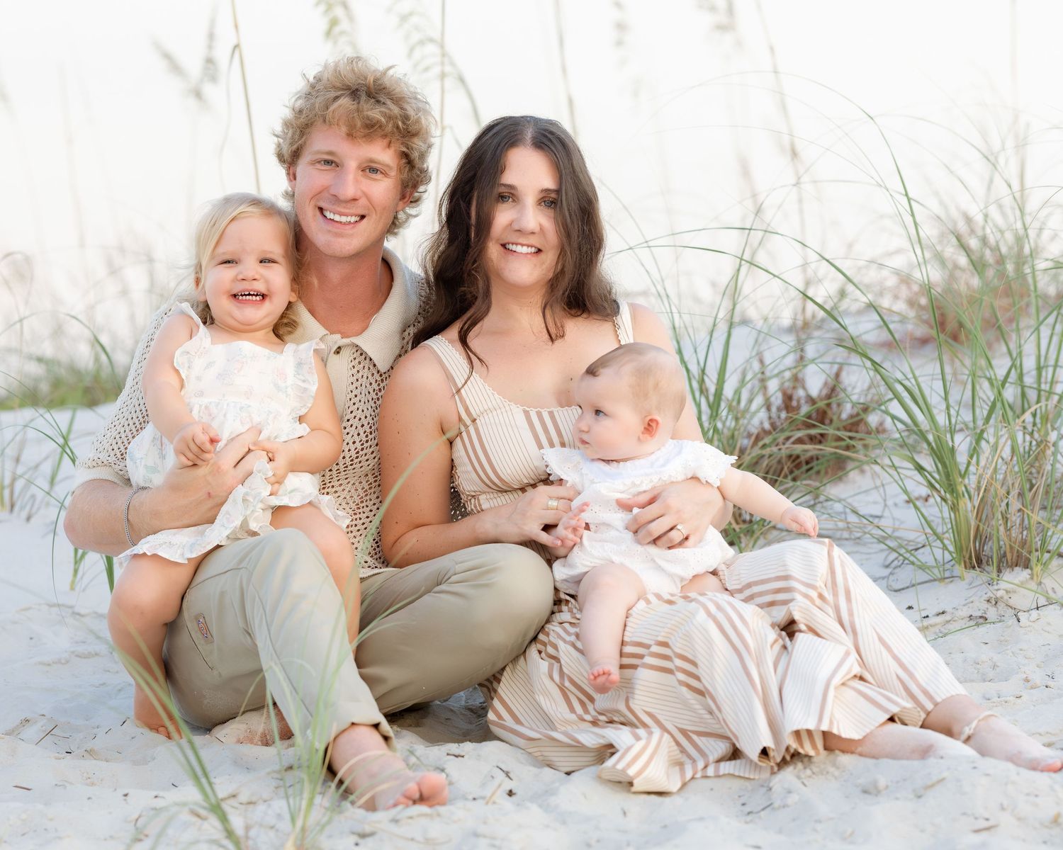 Happy family of four posing on sandy beach, parents smiling with two young children amid coastal dunes.