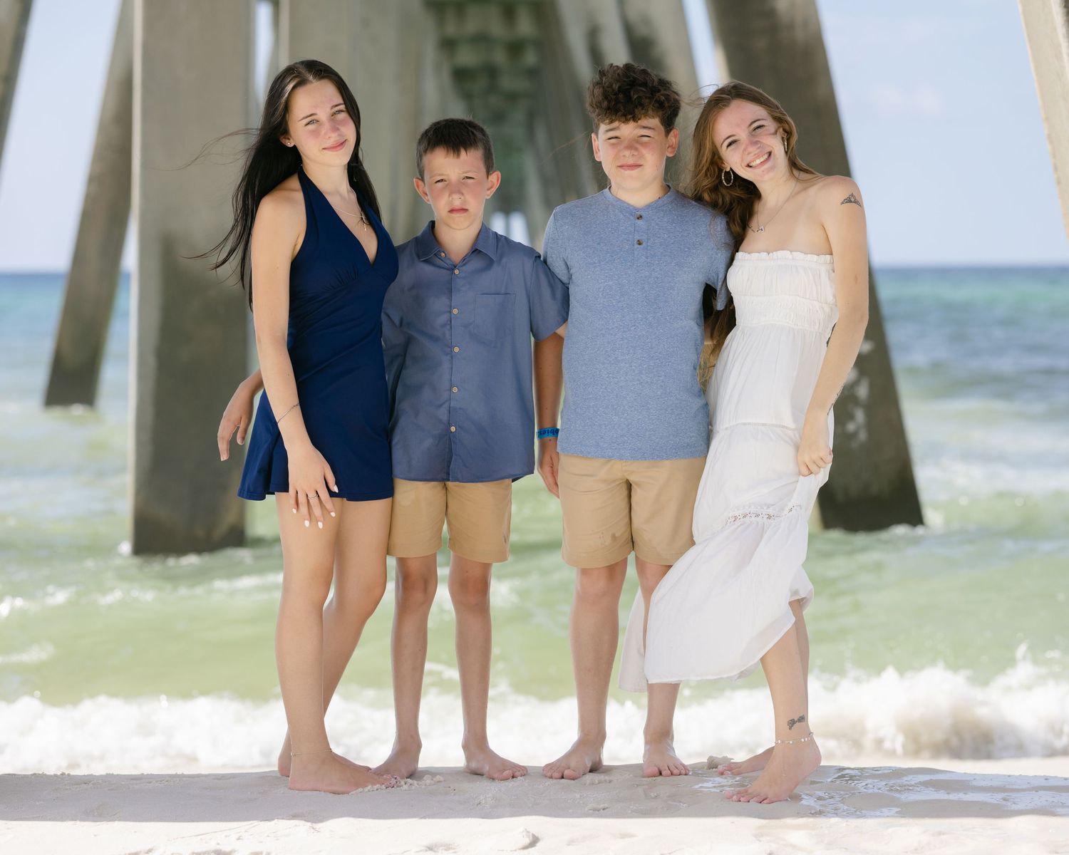 Four teenagers pose barefoot on a sunny beach near a pier with turquoise ocean waves in the background.