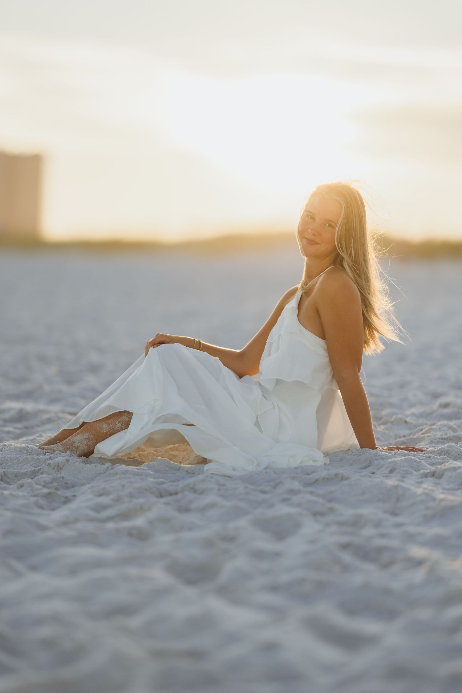 A smiling woman in a white dress sits on a sandy beach at golden sunset, her hair flowing in the breeze.