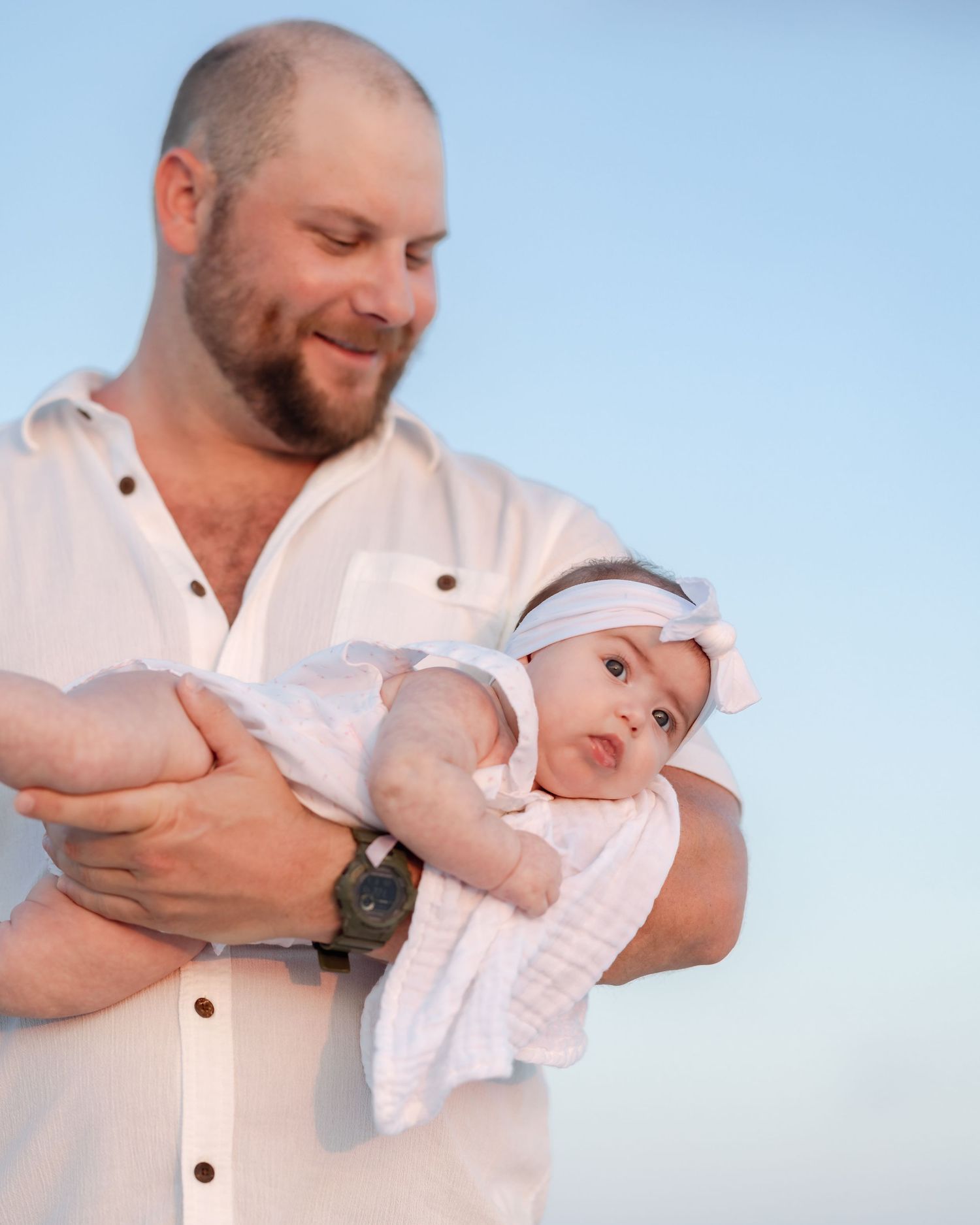 Smiling bearded man in white shirt tenderly holding newborn baby outdoors.