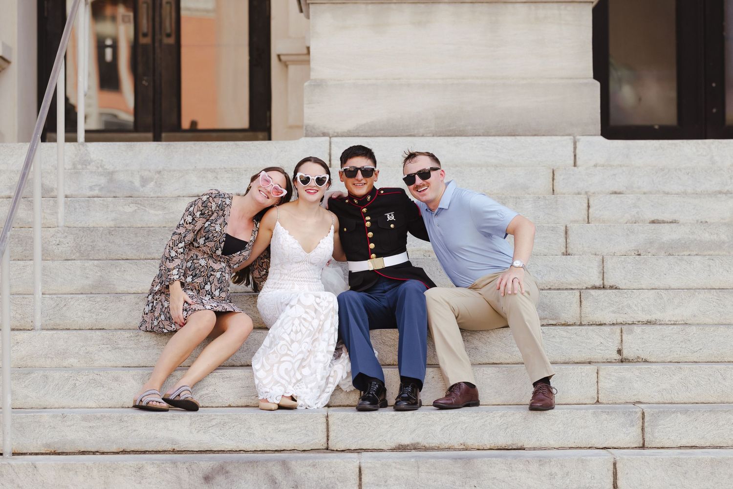 Four friends pose on marble steps, one in Marine dress uniform, smiling together at an outdoor celebration.