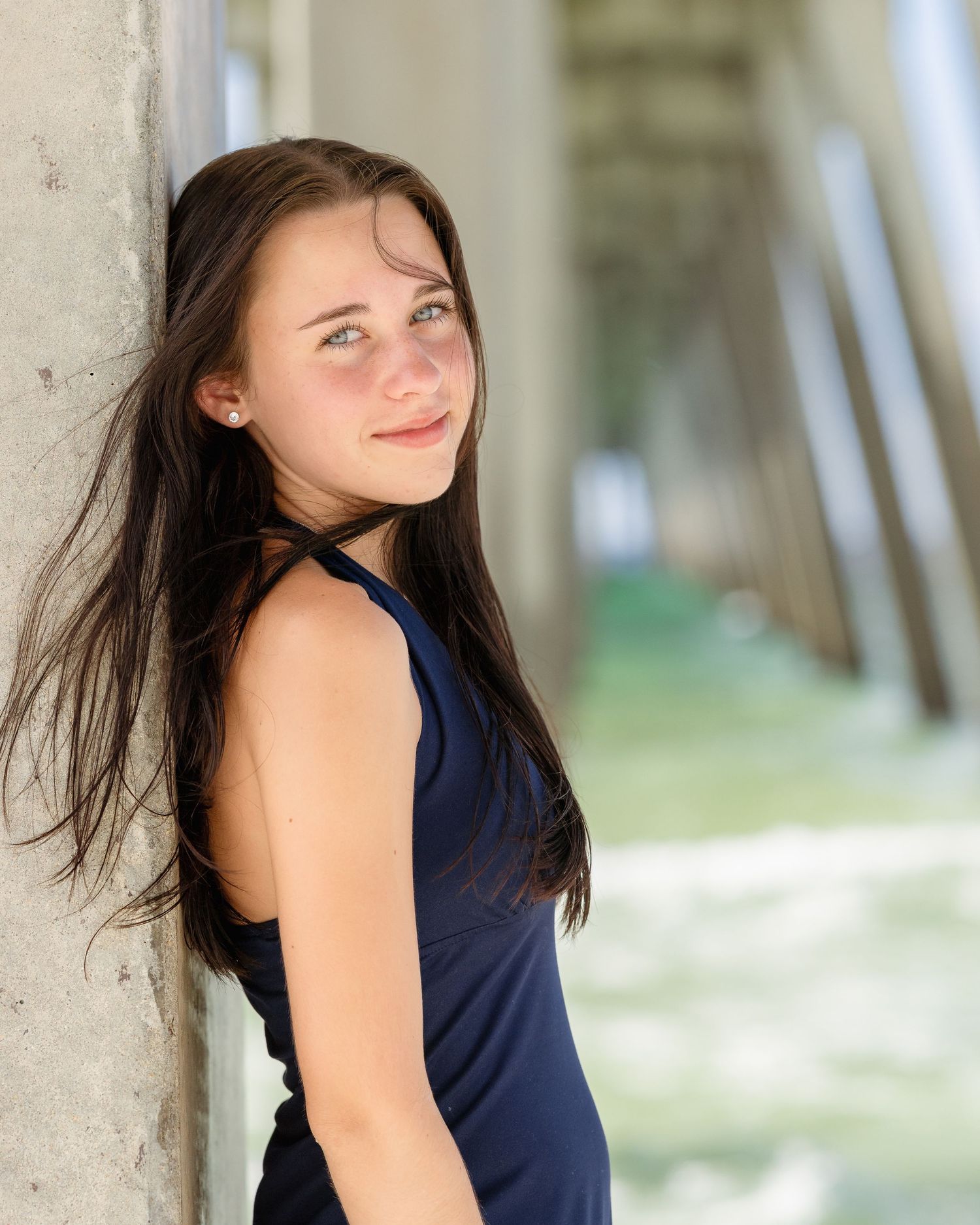Young woman in navy blue top leaning against concrete pillar under beach pier.