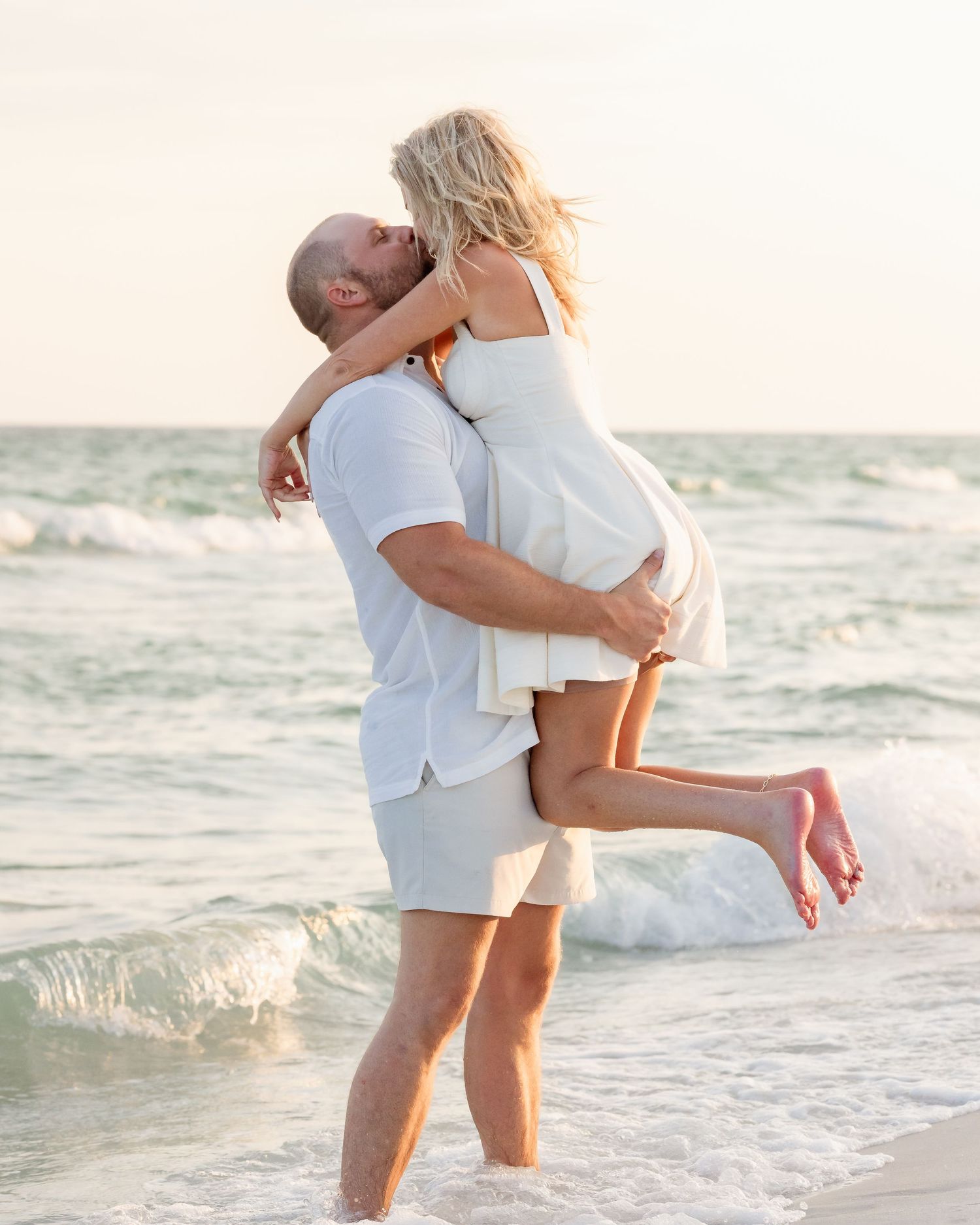 Romantic couple kissing as man lifts woman in white dress at ocean shoreline.