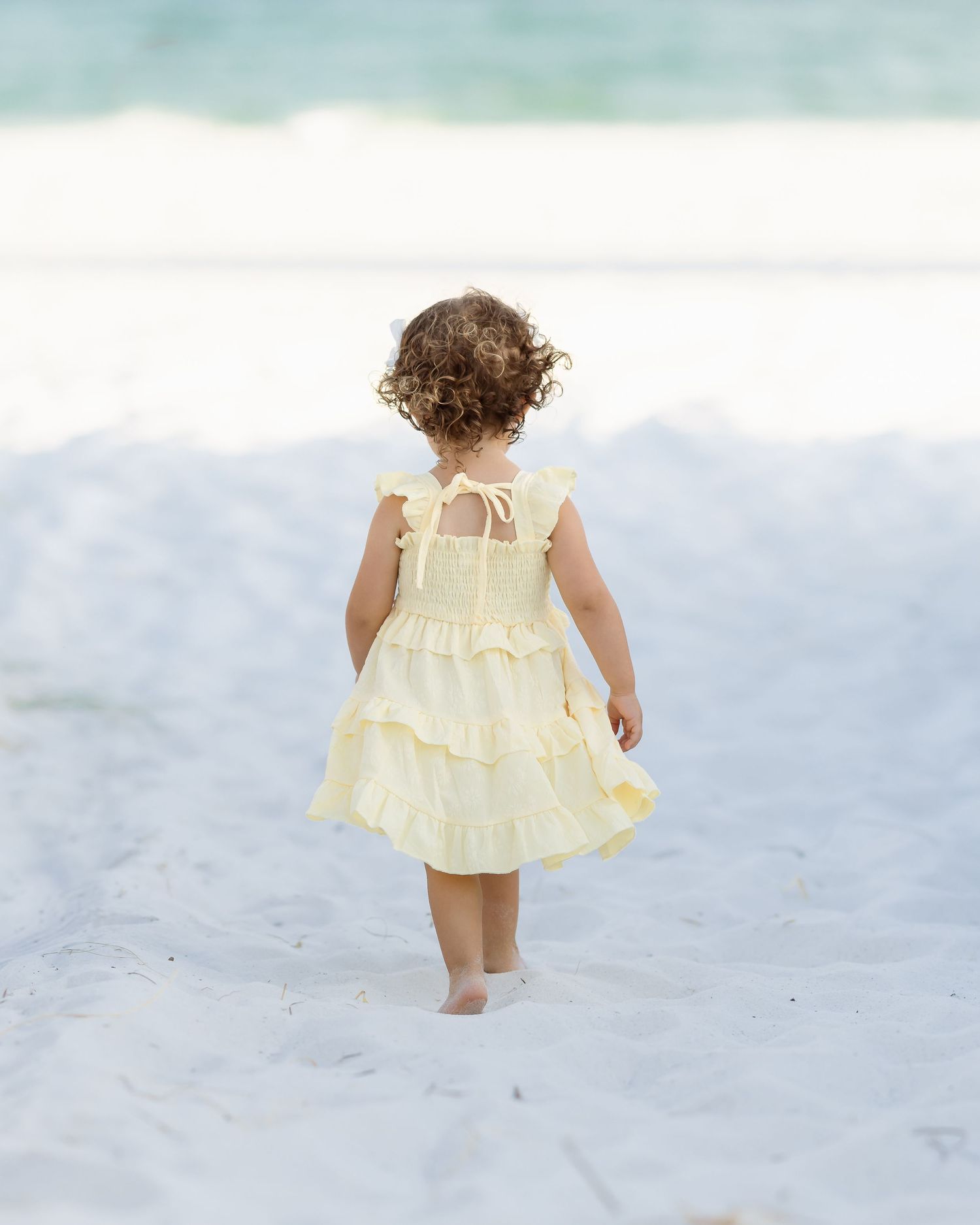 A toddler girl in a yellow ruffled dress walks barefoot on white sand toward ocean waves.