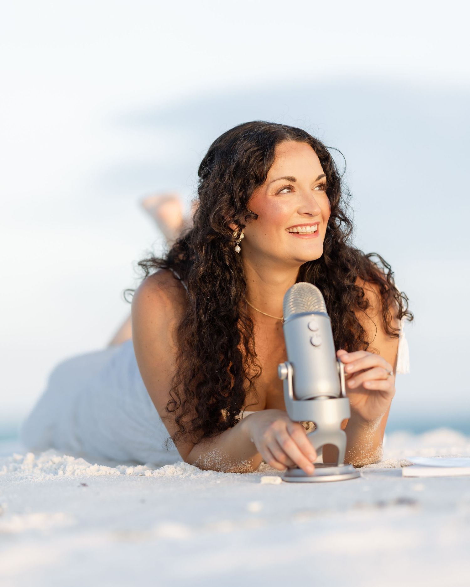 Woman with curly hair lying on white sand beach holding a Blue Yeti microphone, smiling outdoors.