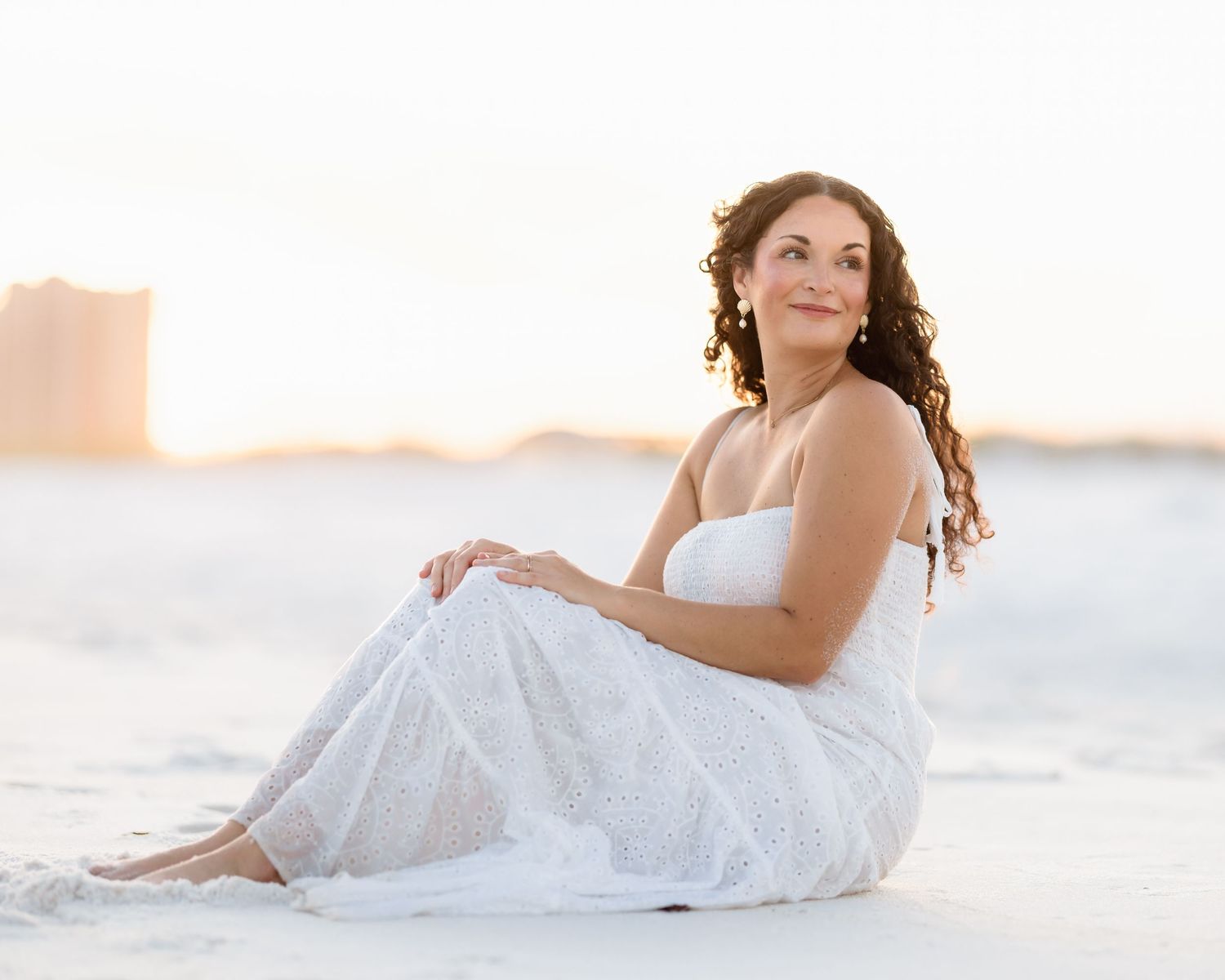 Woman in white dress sitting on sandy beach at golden sunset.