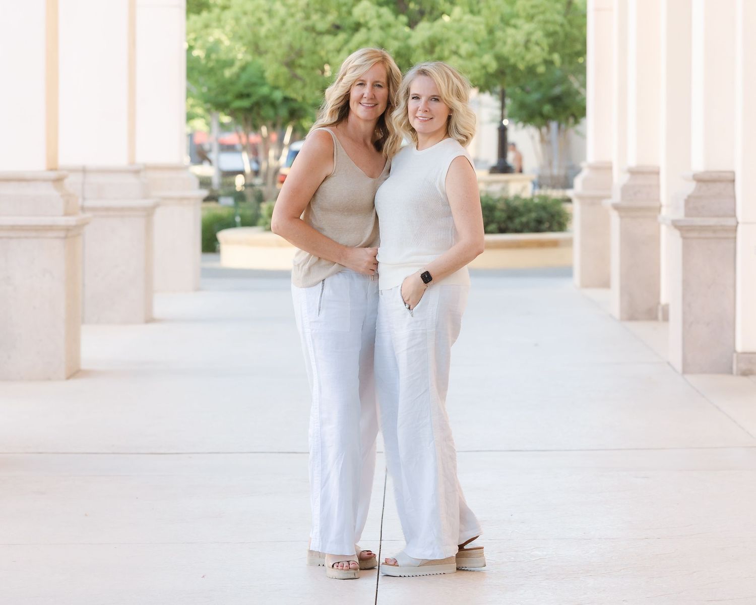Two smiling blonde women in white pants pose together outdoors near elegant columns in a bright, airy setting.