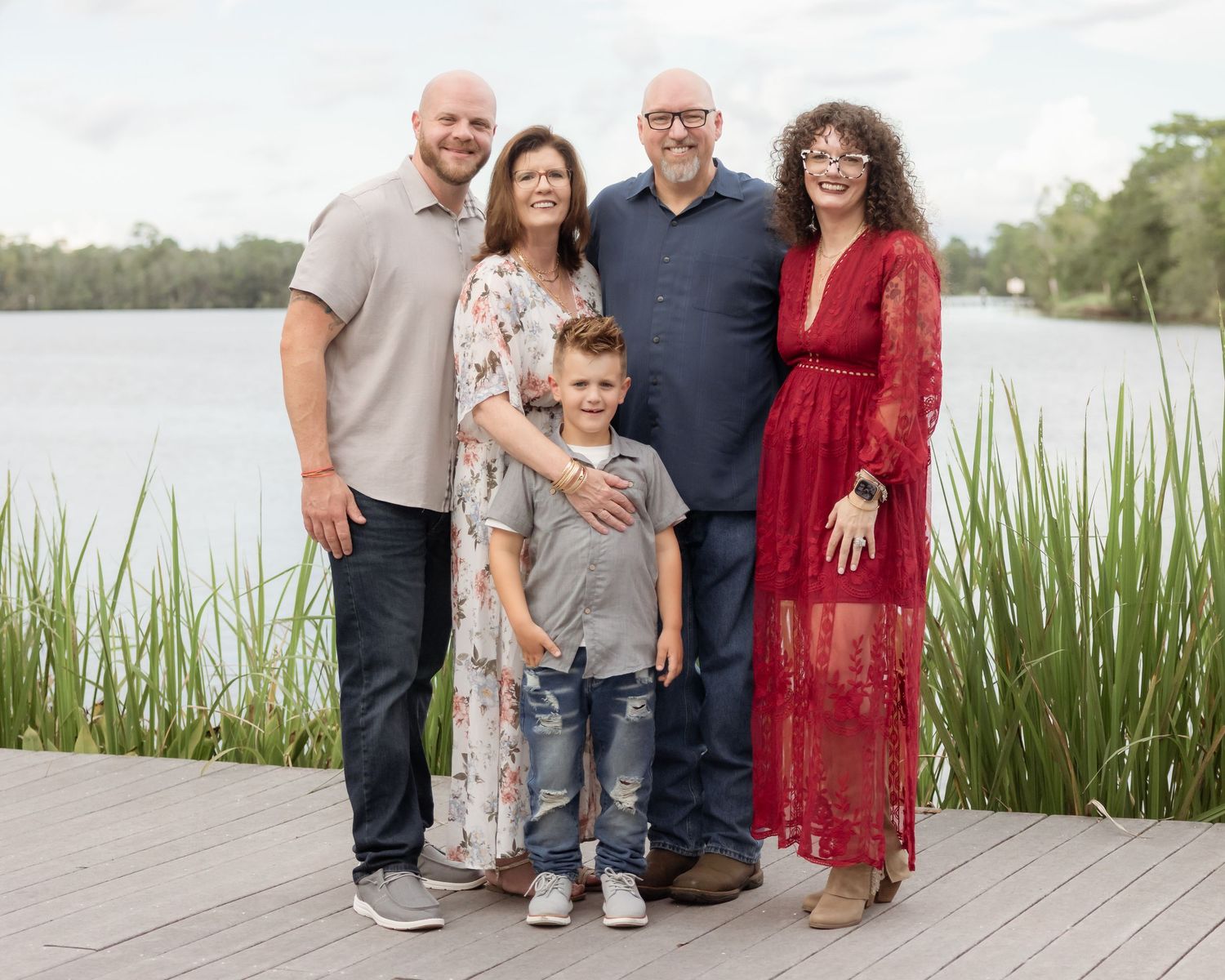 Family of five posing outdoors on a wooden deck near a serene lake surrounded by lush greenery.