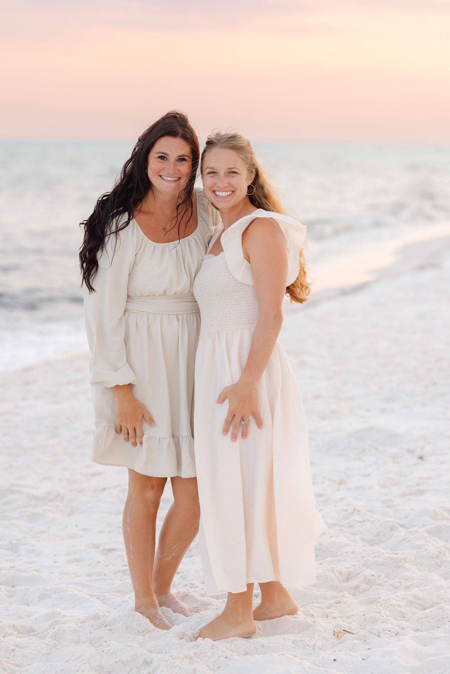 Mother and daughter wearing white dresses enjoying a tender moment together on a sandy beach at sunset.