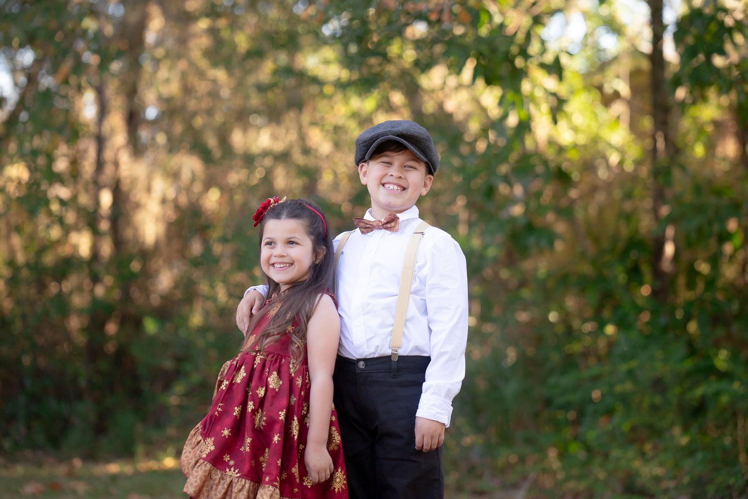 Two young children in formal attire standing together outdoors with a wooded background in soft lighting.