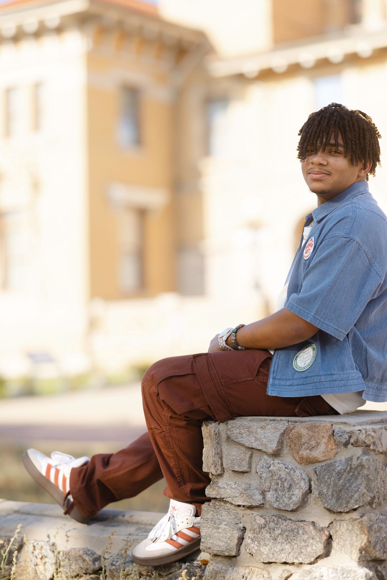 Young man with dreadlocks sitting on stone wall wearing blue shirt, brown cargo pants, and Adidas sneakers outdoors.