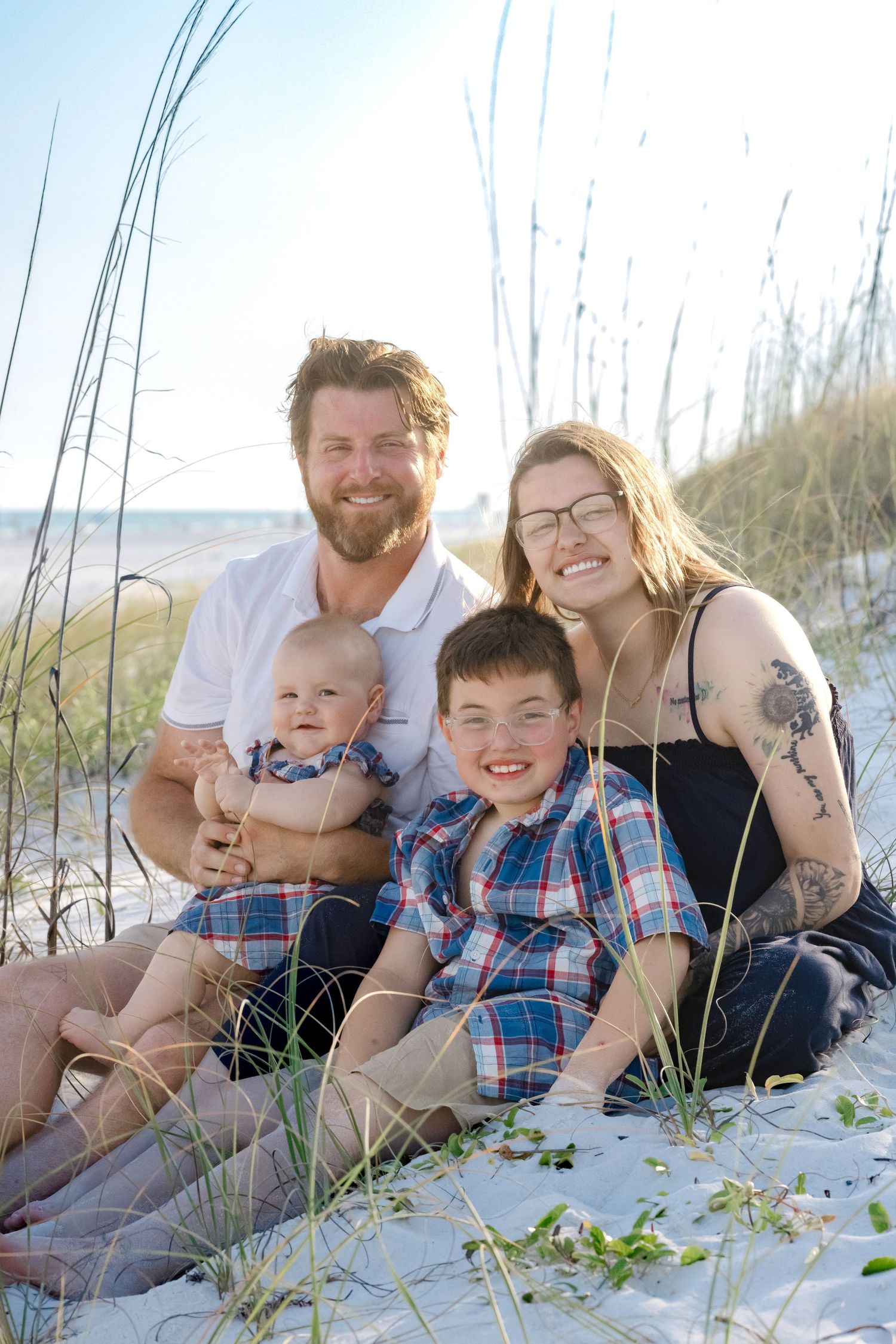 Family of four smiling together on sandy beach dunes with sea grass, parents holding baby and young boy.