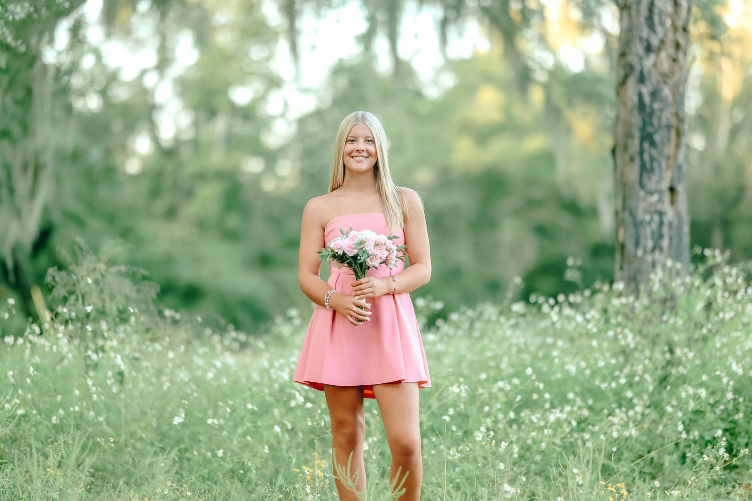 Young blonde woman in pink dress holding bouquet in sunny green meadow with trees.