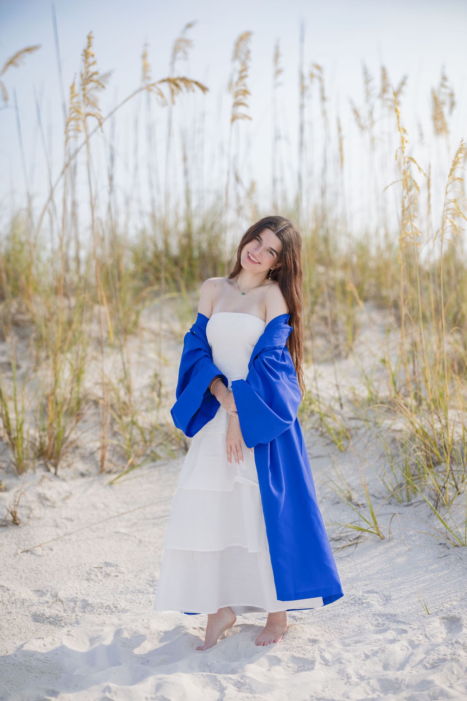 Smiling young woman in white dress with blue coat posing barefoot on sandy beach dunes.