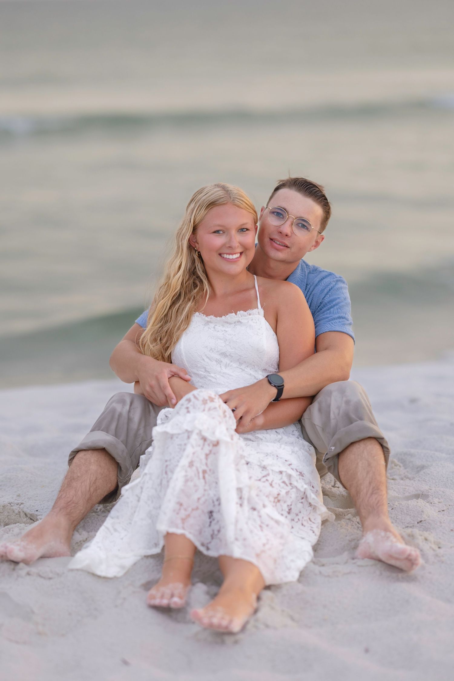 Couple sitting together on sandy beach, woman in white lace dress, man in blue shirt, smiling at camera near ocean.