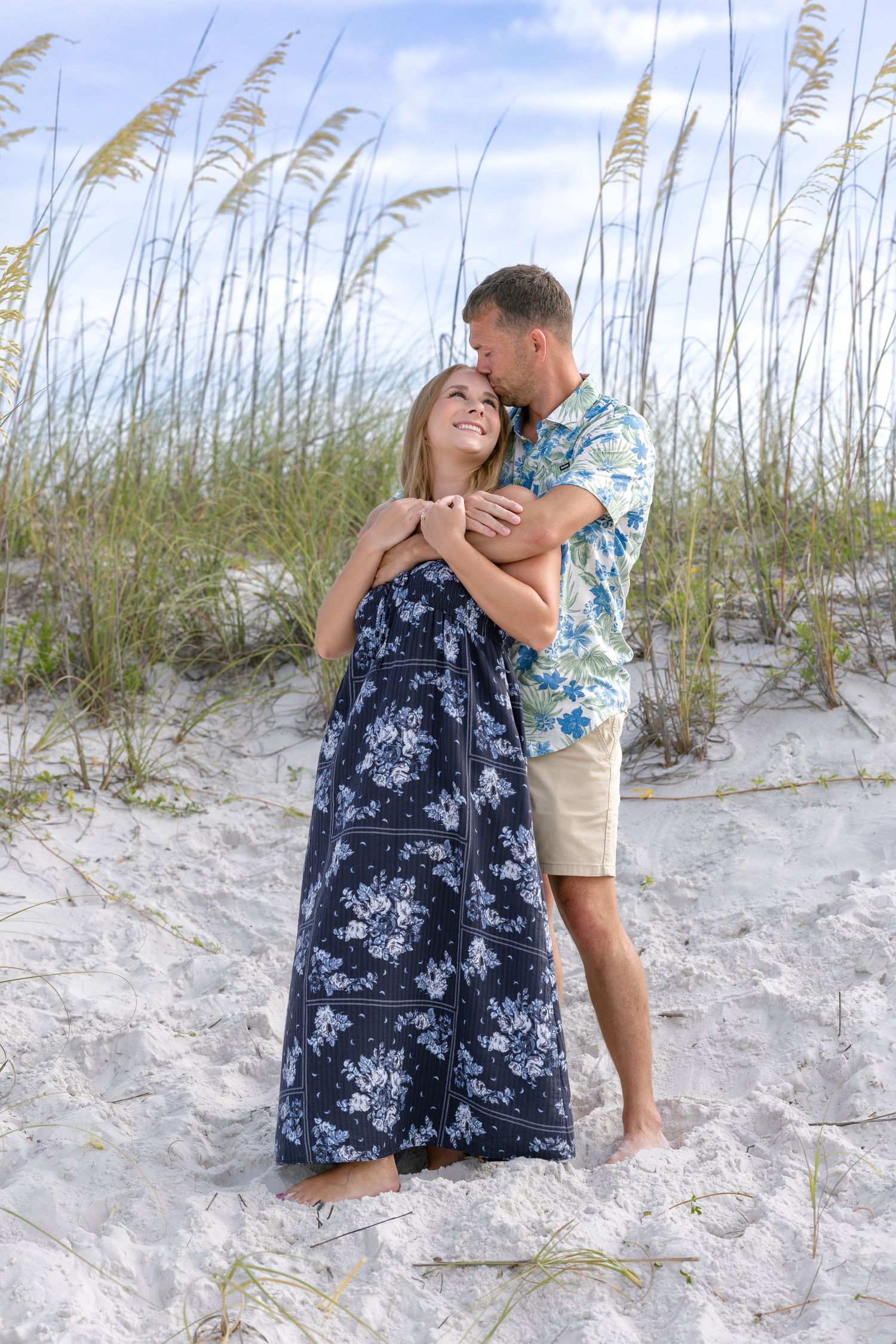 A romantic couple embraces on white sand beach dunes with sea oats, woman in floral dress, man in Hawaiian shirt.