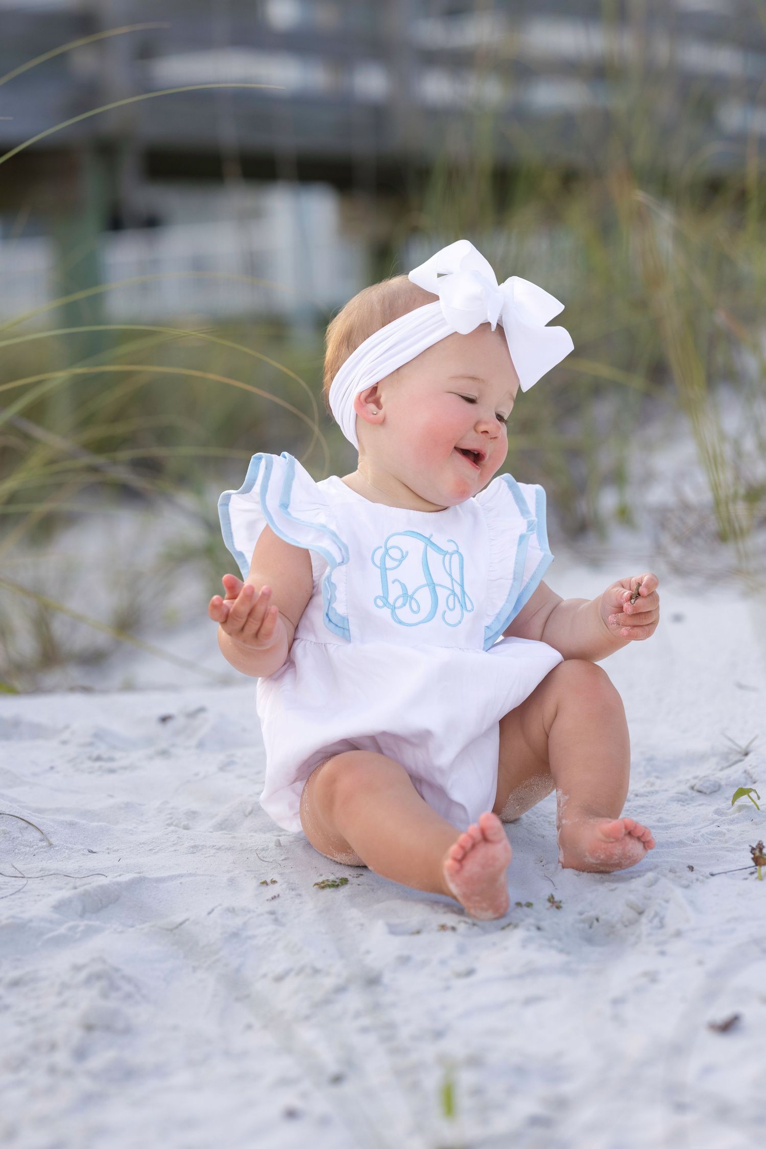 Happy baby girl in white monogrammed romper and bow headband laughing on white sand beach.