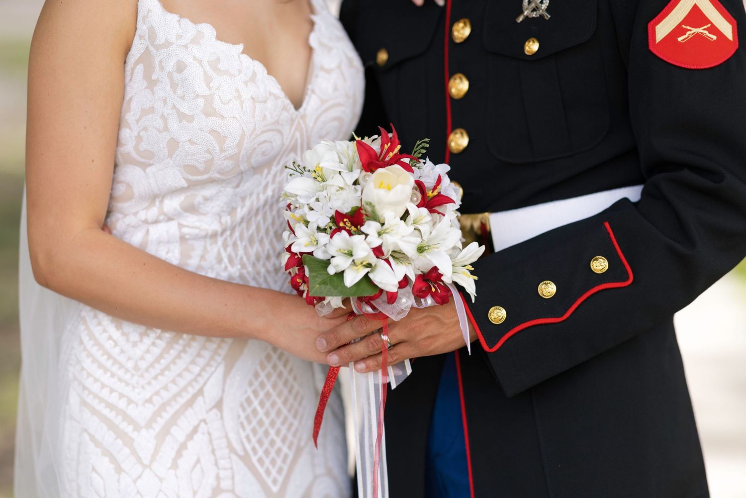 Marine in dress uniform and bride in white lace gown holding red and white wedding bouquet together.