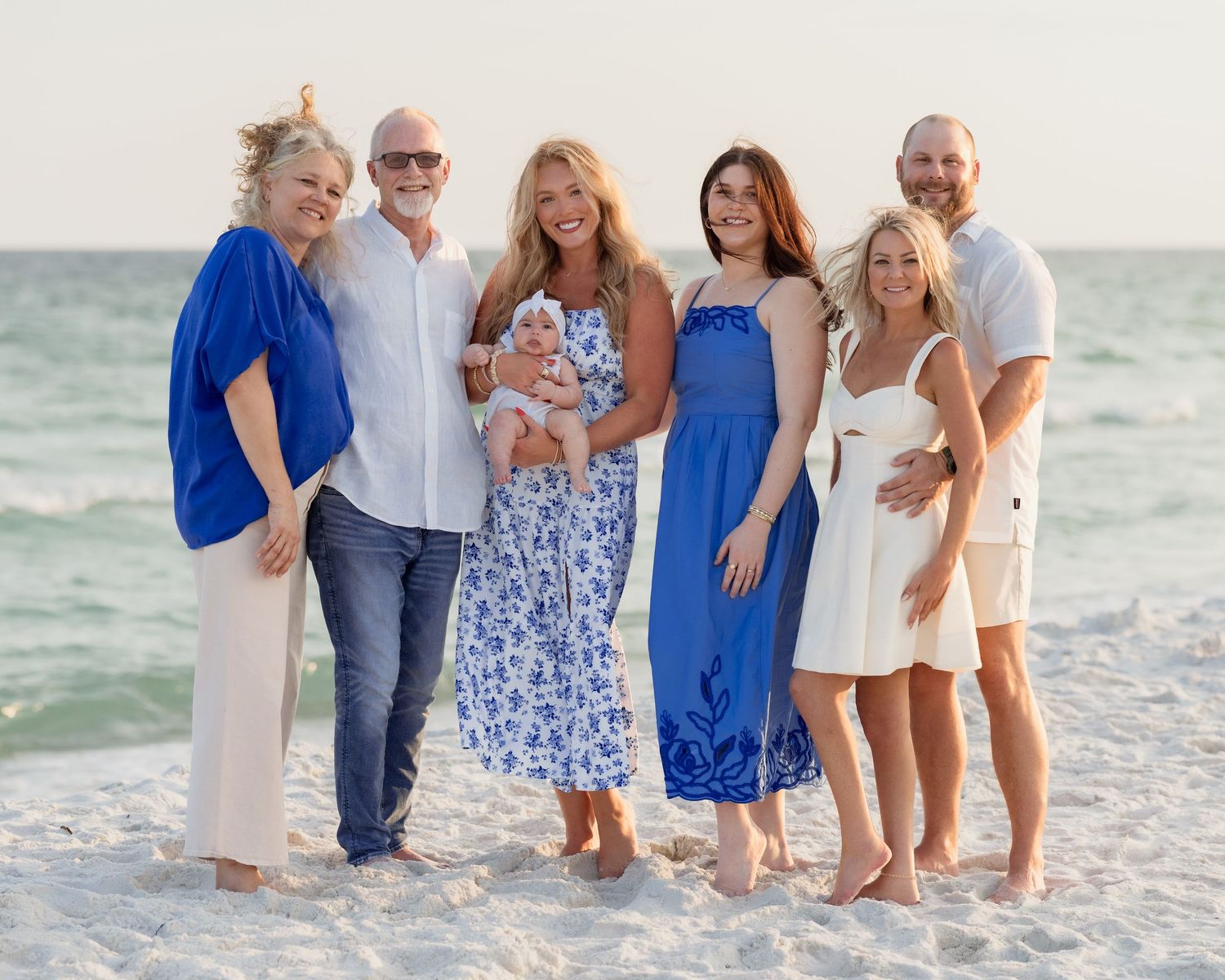 Family of seven posing together on a sandy beach at sunset, wearing coordinating blue and white outfits.