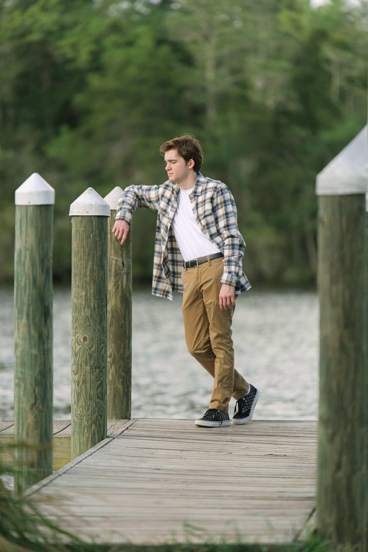 Young man in plaid shirt and khakis leaning on dock posts by calm water with lush green trees in background.