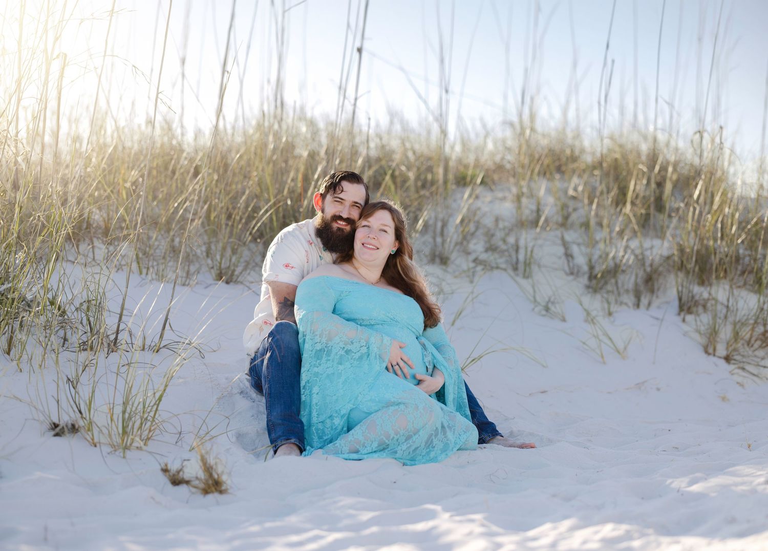Pregnant couple sitting together on white sand beach dunes, woman in teal lace maternity dress, man in white shirt.