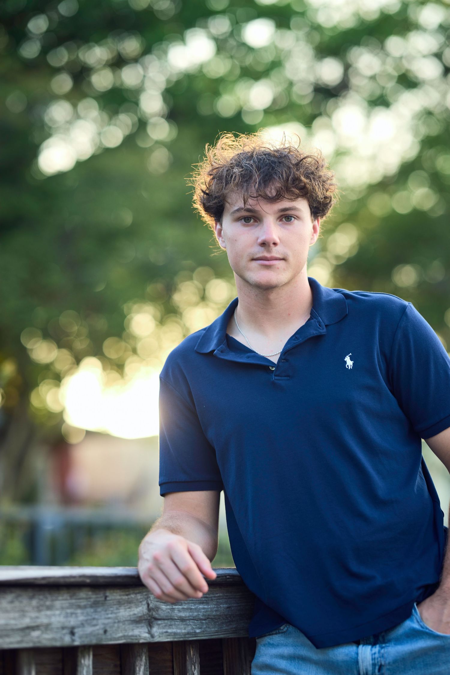 Young man with curly hair in navy Ralph Lauren polo leans on wooden fence outdoors with bokeh green trees background.
