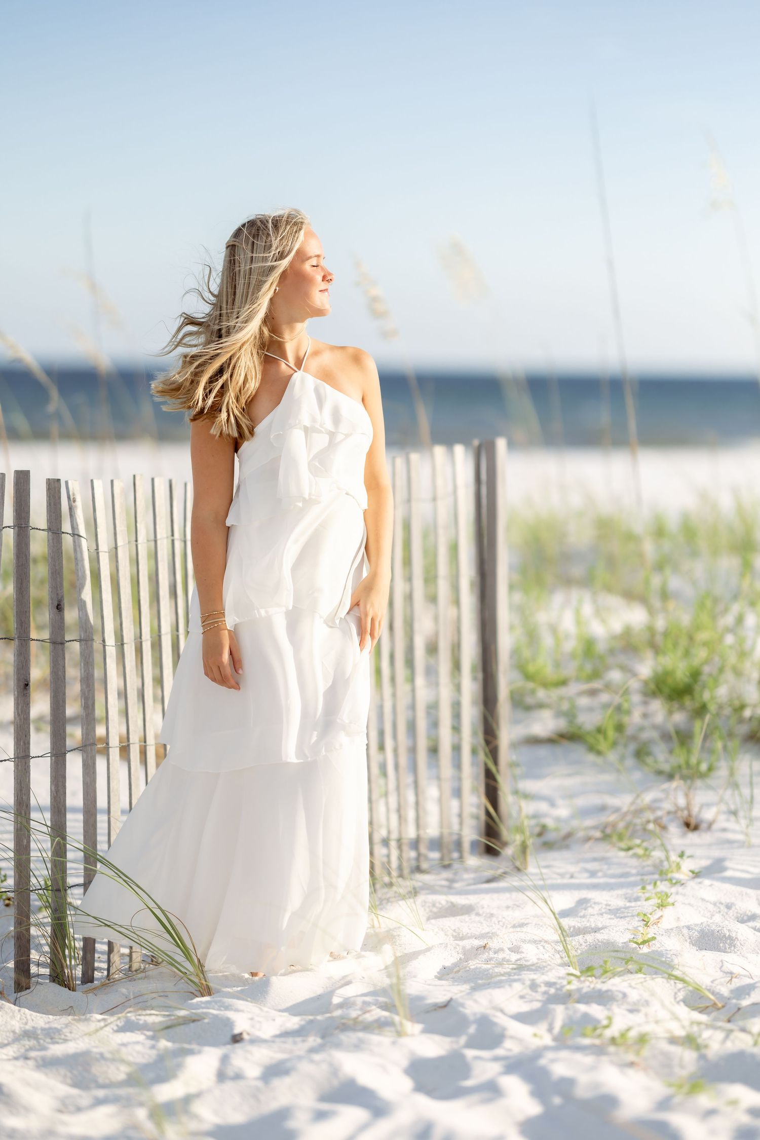 Woman in white maxi dress standing by beach fence with sea oats, smiling in golden sunlight.