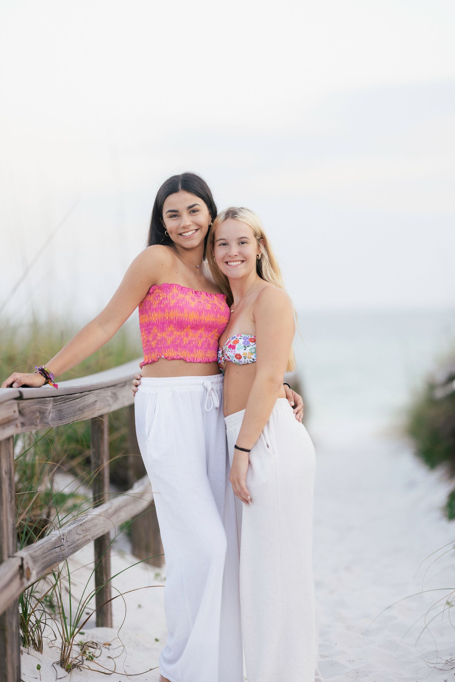 Two people in white outfits pose together on sandy beach boardwalk at sunset.
