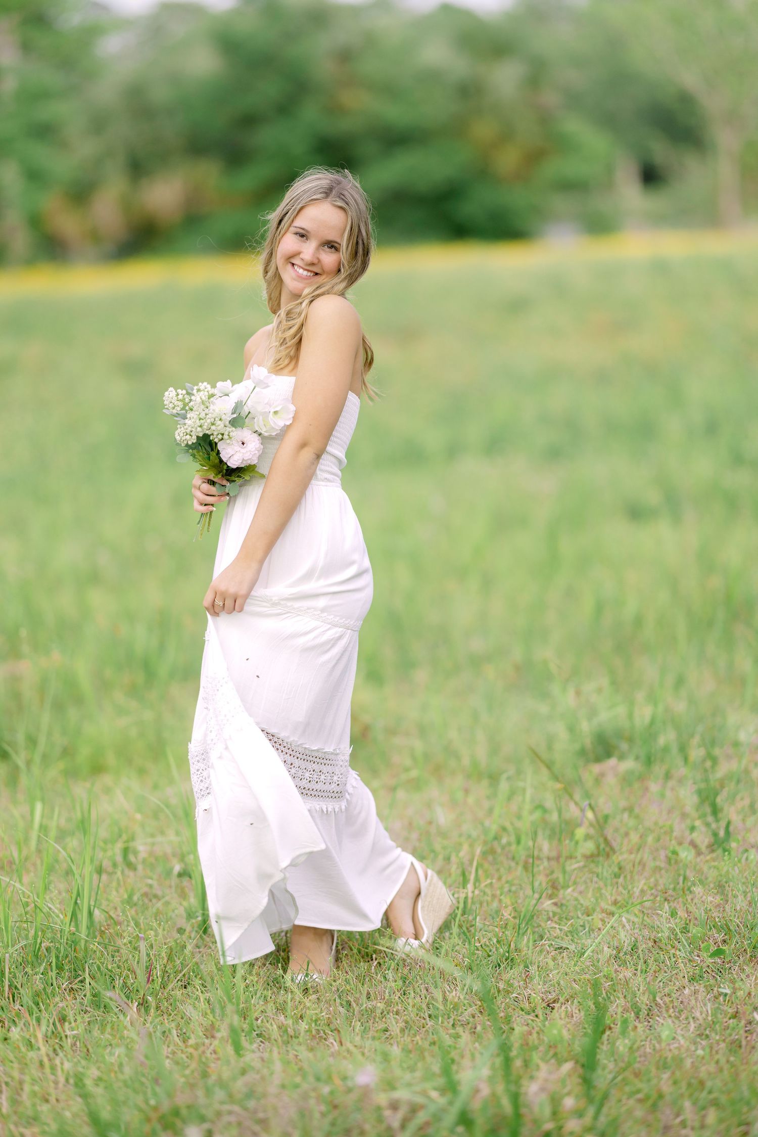 A person in a flowing white summer dress holding a bouquet of white flowers stands in a sunlit meadow.