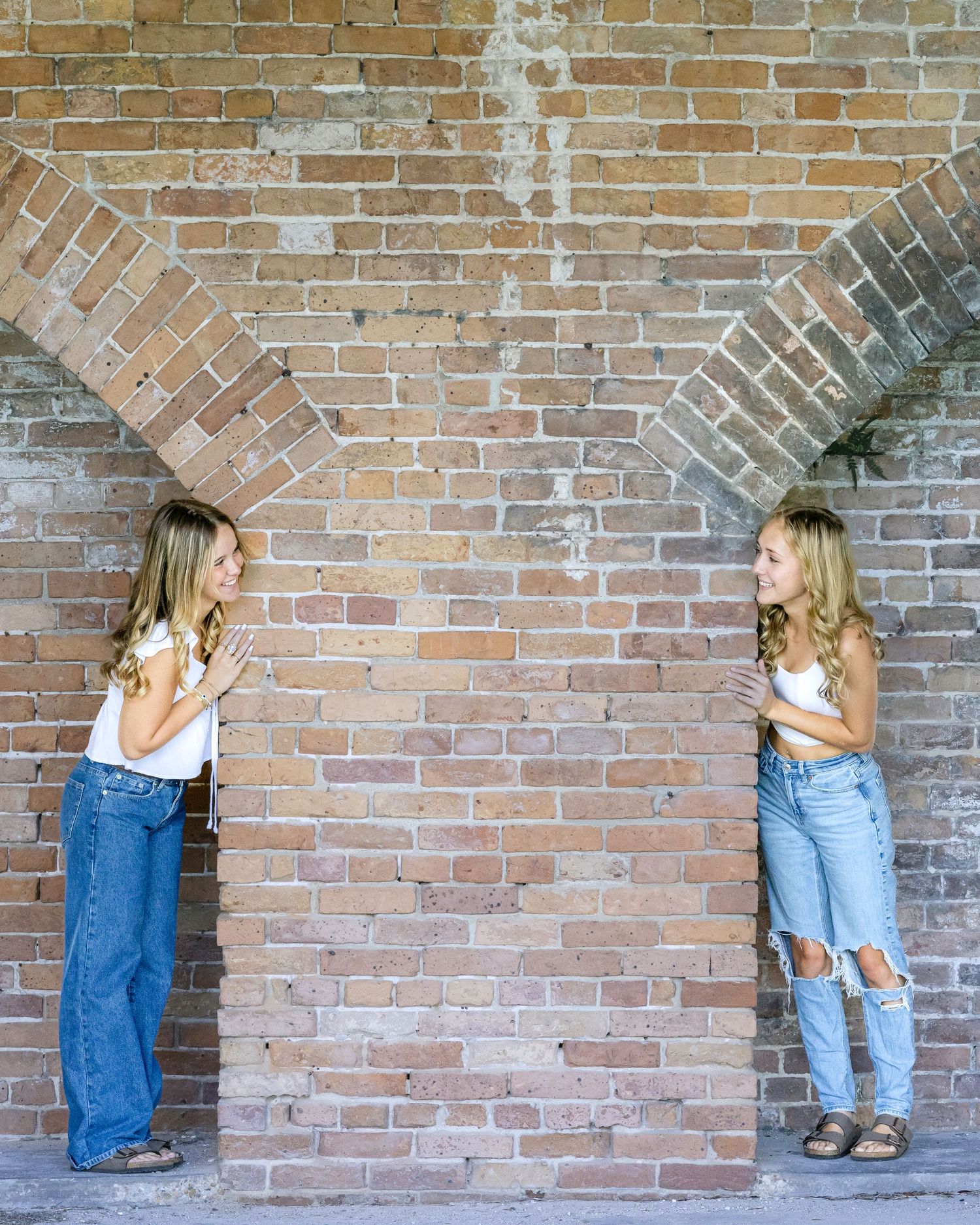 Two people stand in opposite brick arches along a weathered wall, wearing casual denim outfits in contrasting poses.