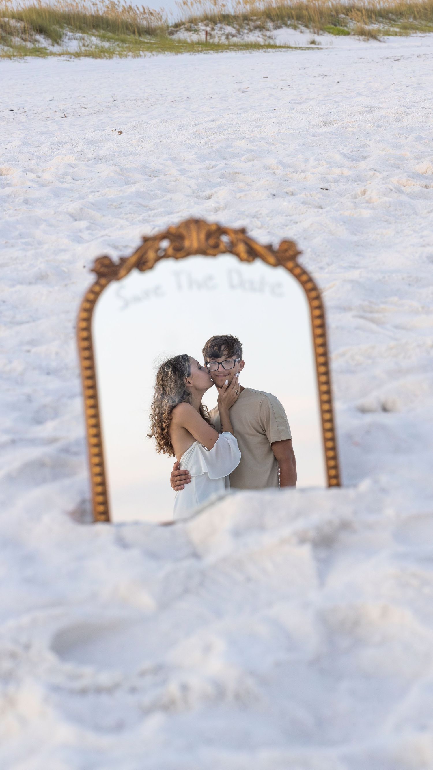 An ornate gold mirror reflects a romantic scene while placed in white sand on a beach.