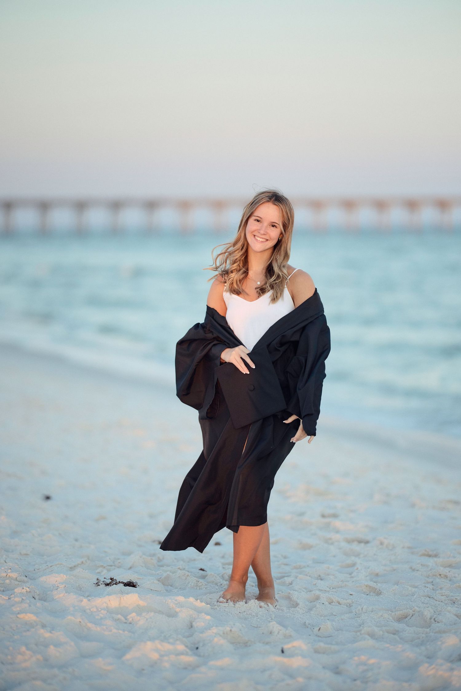 A person in a black outfit poses on a sandy beach at sunset with turquoise water in the background.