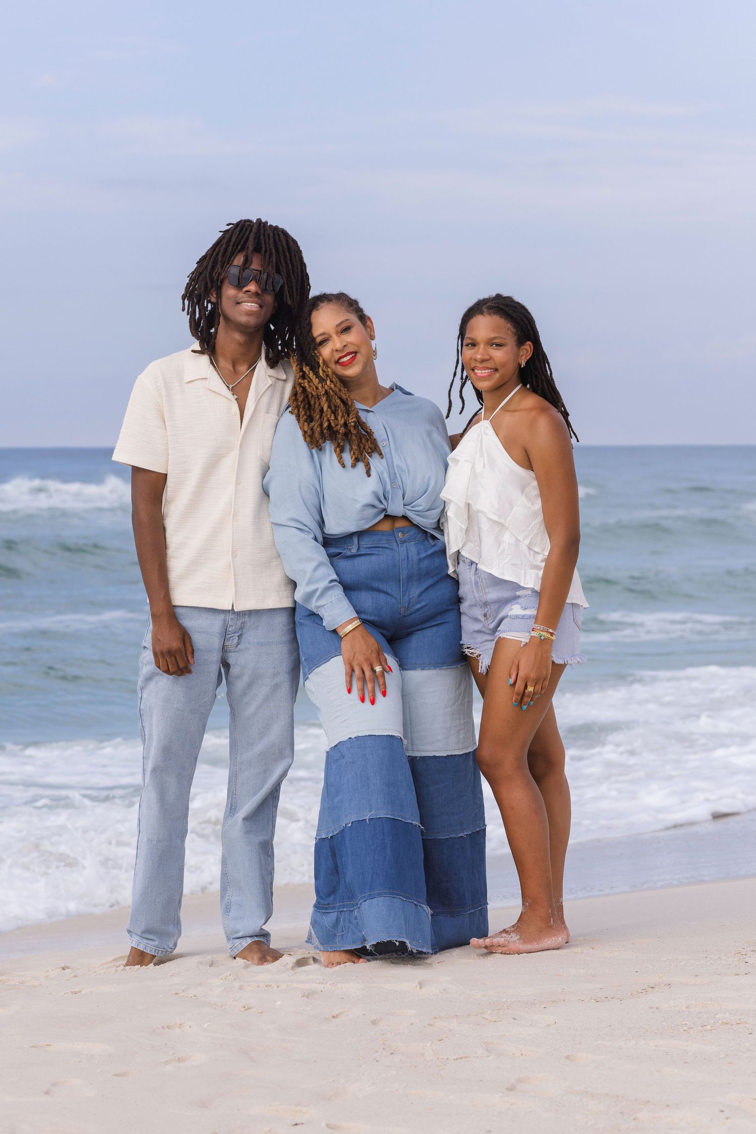 Three young Black adults pose together on a sandy beach, dressed in coordinating blue and white casual outfits.