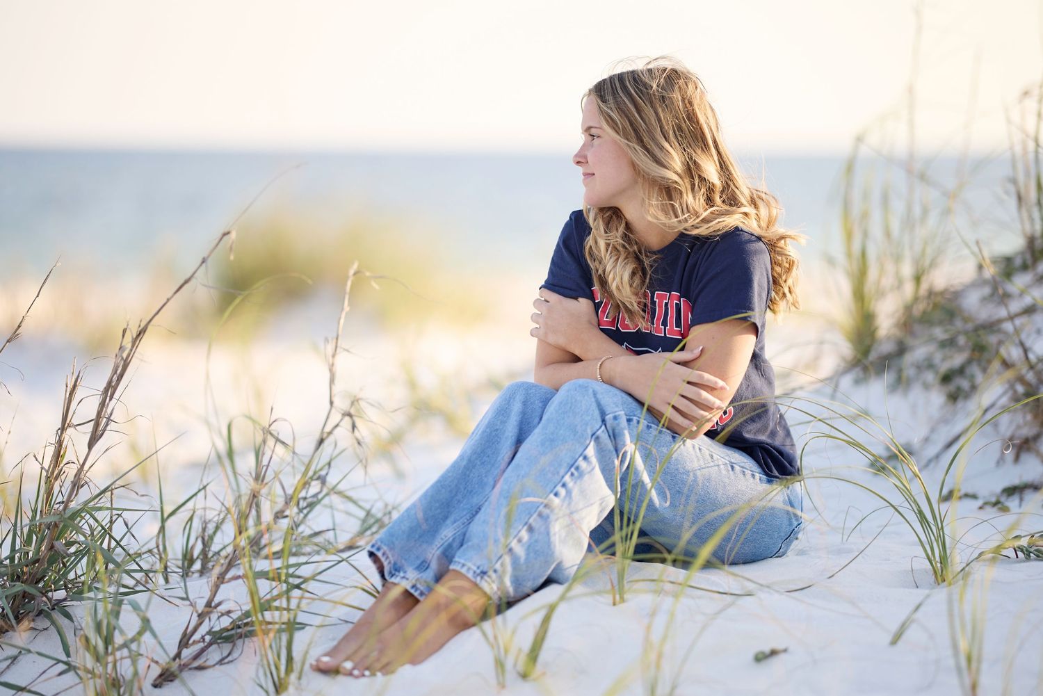 Young woman sitting on sandy beach dunes, wearing jeans and navy shirt, gazing at the ocean on a sunny day.