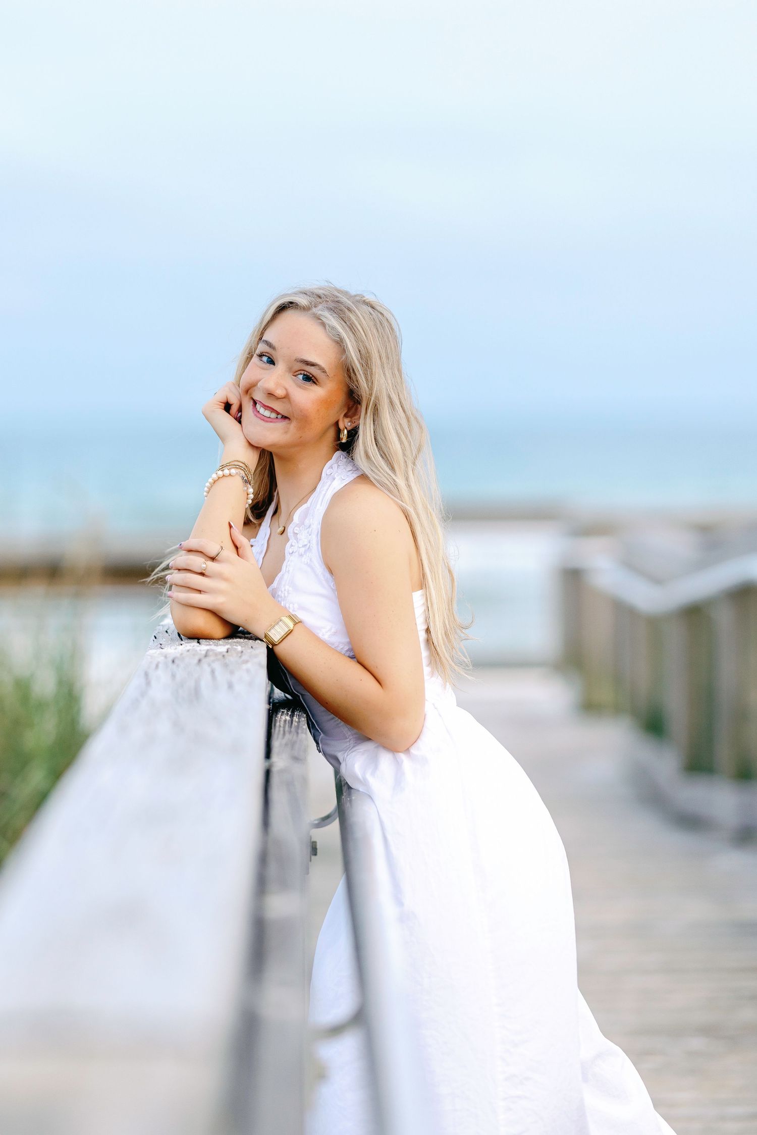 Smiling blonde woman in white dress leaning on wooden boardwalk railing near beach on a clear day.