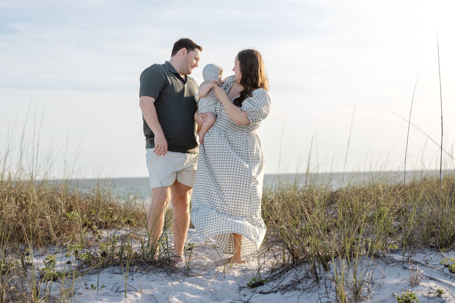 Family of three poses on sandy beach dunes with sea oats, parents gazing at newborn baby in soft natural light.