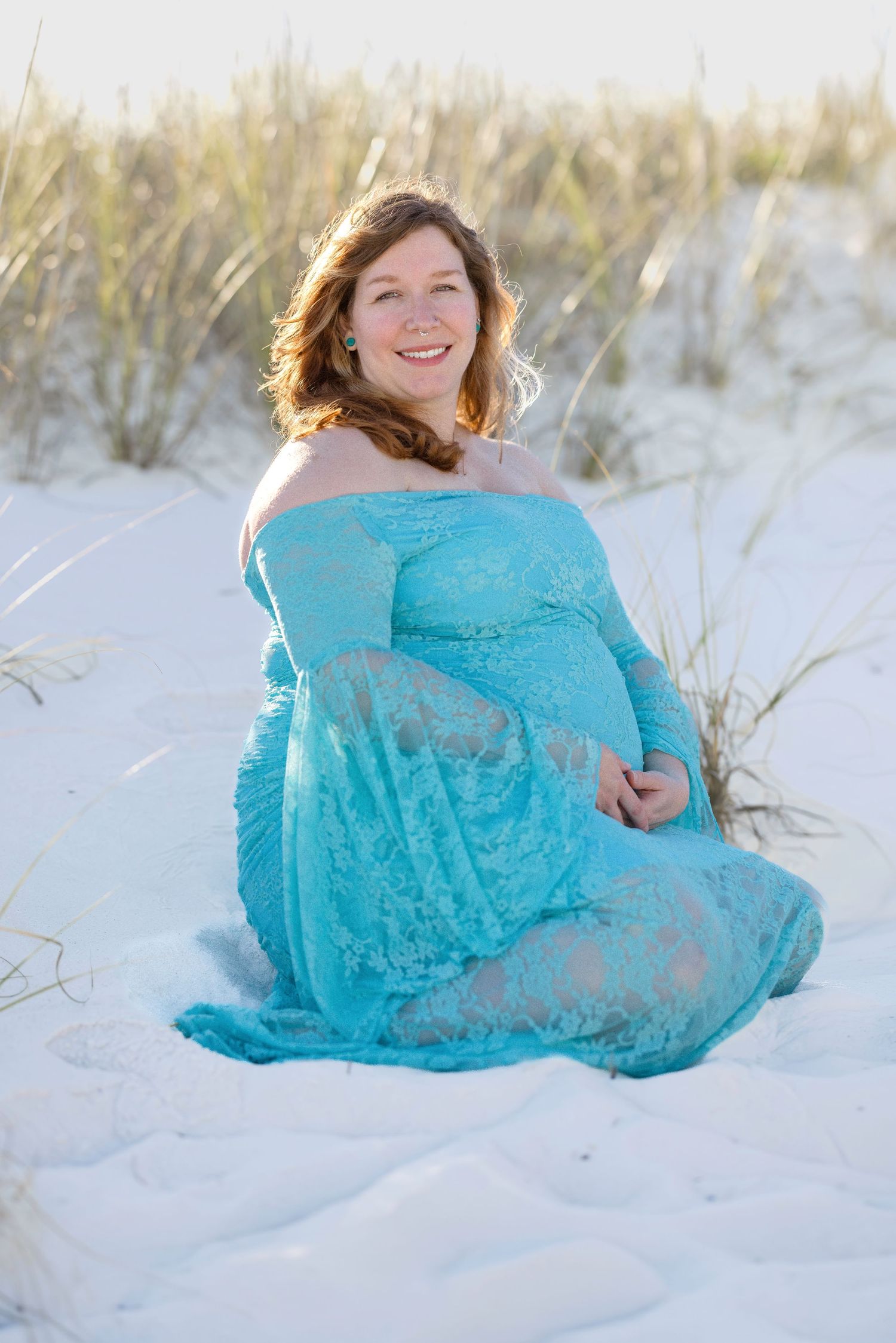 Pregnant woman in turquoise lace off-shoulder gown sitting on white sand beach dunes, smiling warmly.