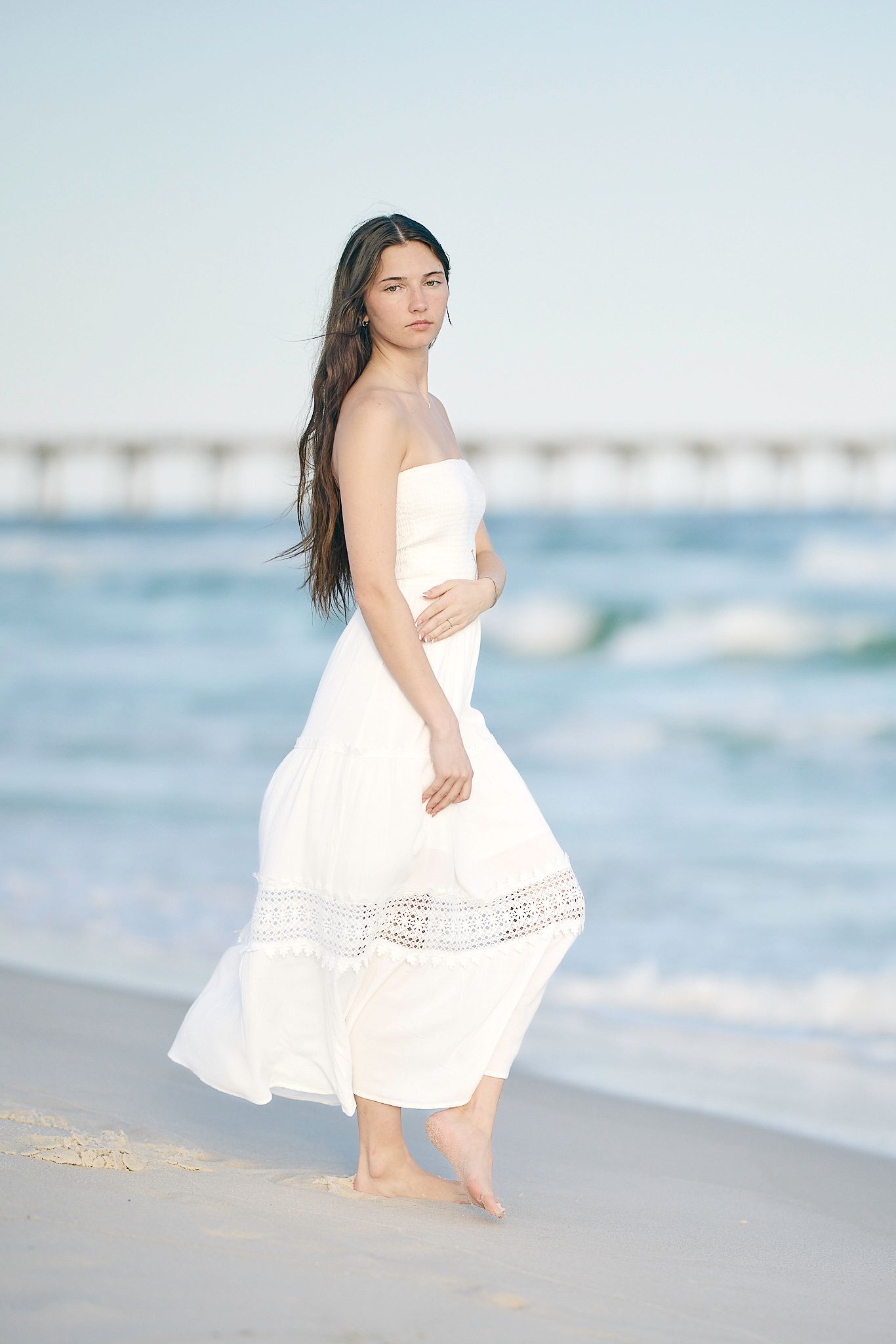 Young woman in white strapless maxi dress standing barefoot on sandy beach with ocean waves and pier in background.