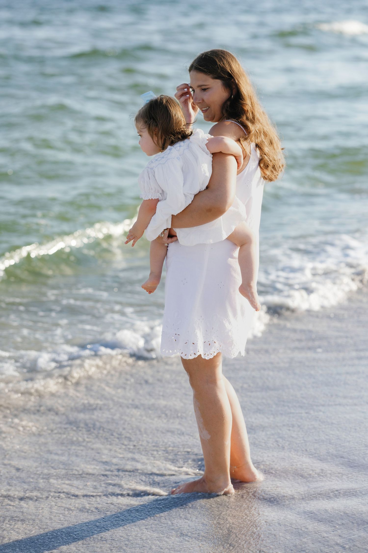 A mother in a white dress holds her baby girl while standing barefoot on a sunny beach with ocean waves.