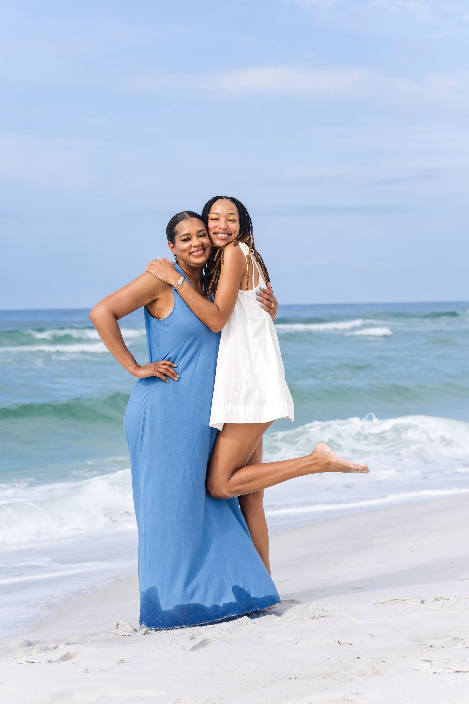 Two women hugging and smiling on a sunny beach, one in a blue maxi dress and one in a white dress.