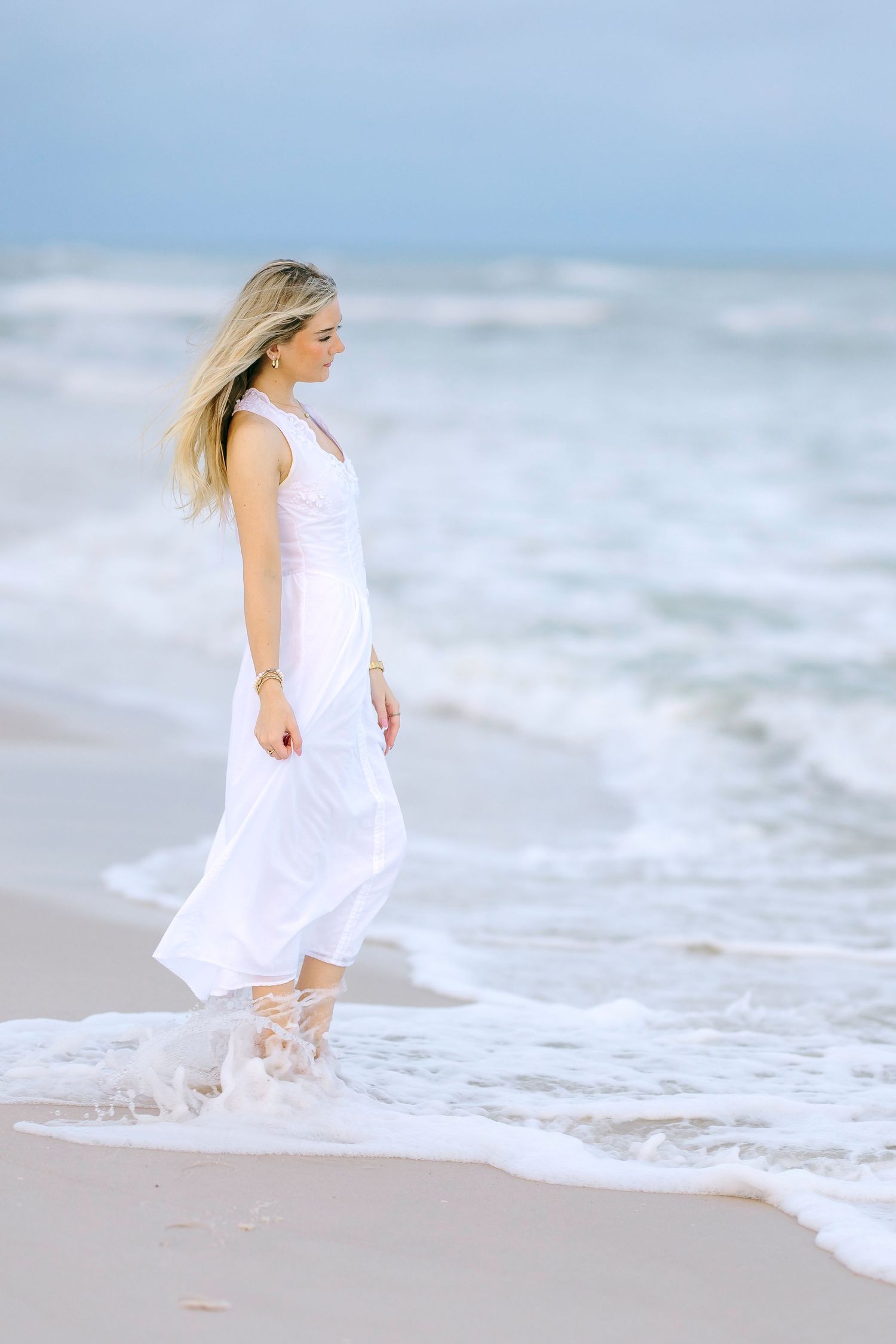 Blonde woman in flowing white maxi dress standing in ocean waves on a sandy beach.