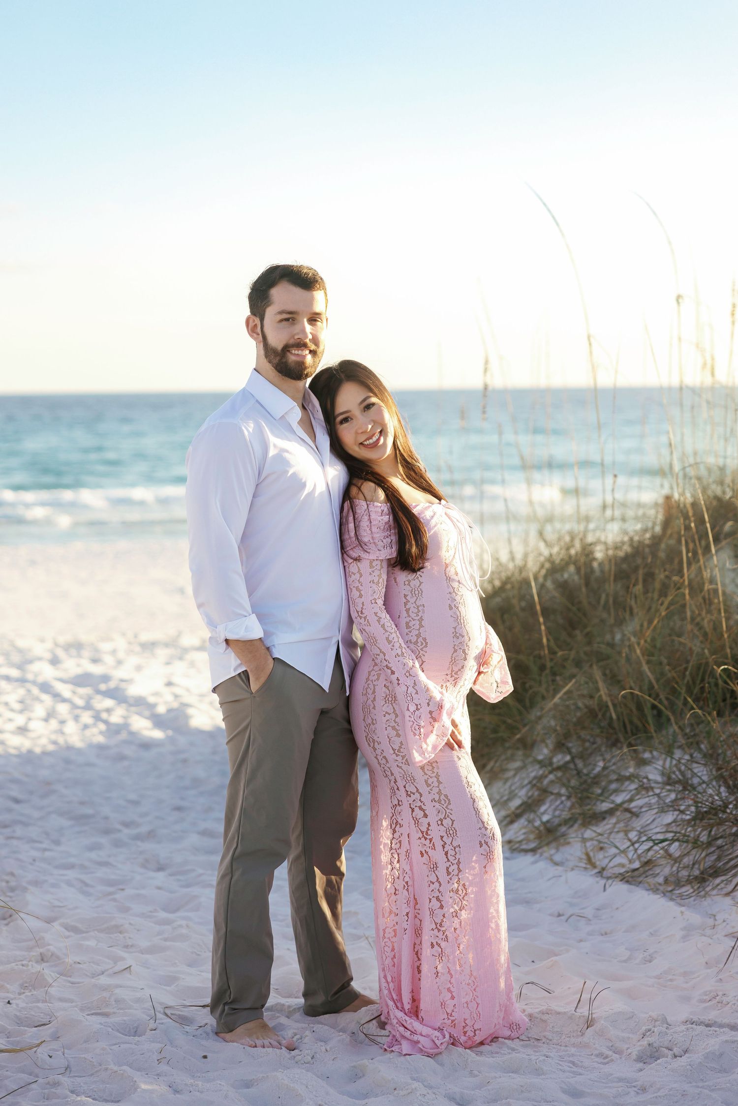 Couple posing on a sandy beach, woman in pink lace dress, man in white shirt, with ocean and dunes in background.